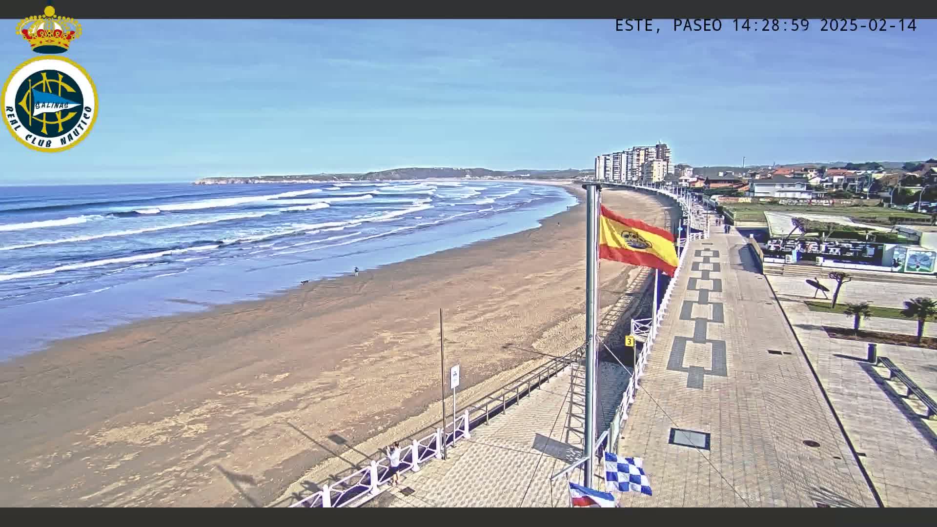 A sandy beach with waves rolling in under a clear blue sky is viewed from a paved walkway alongside buildings.