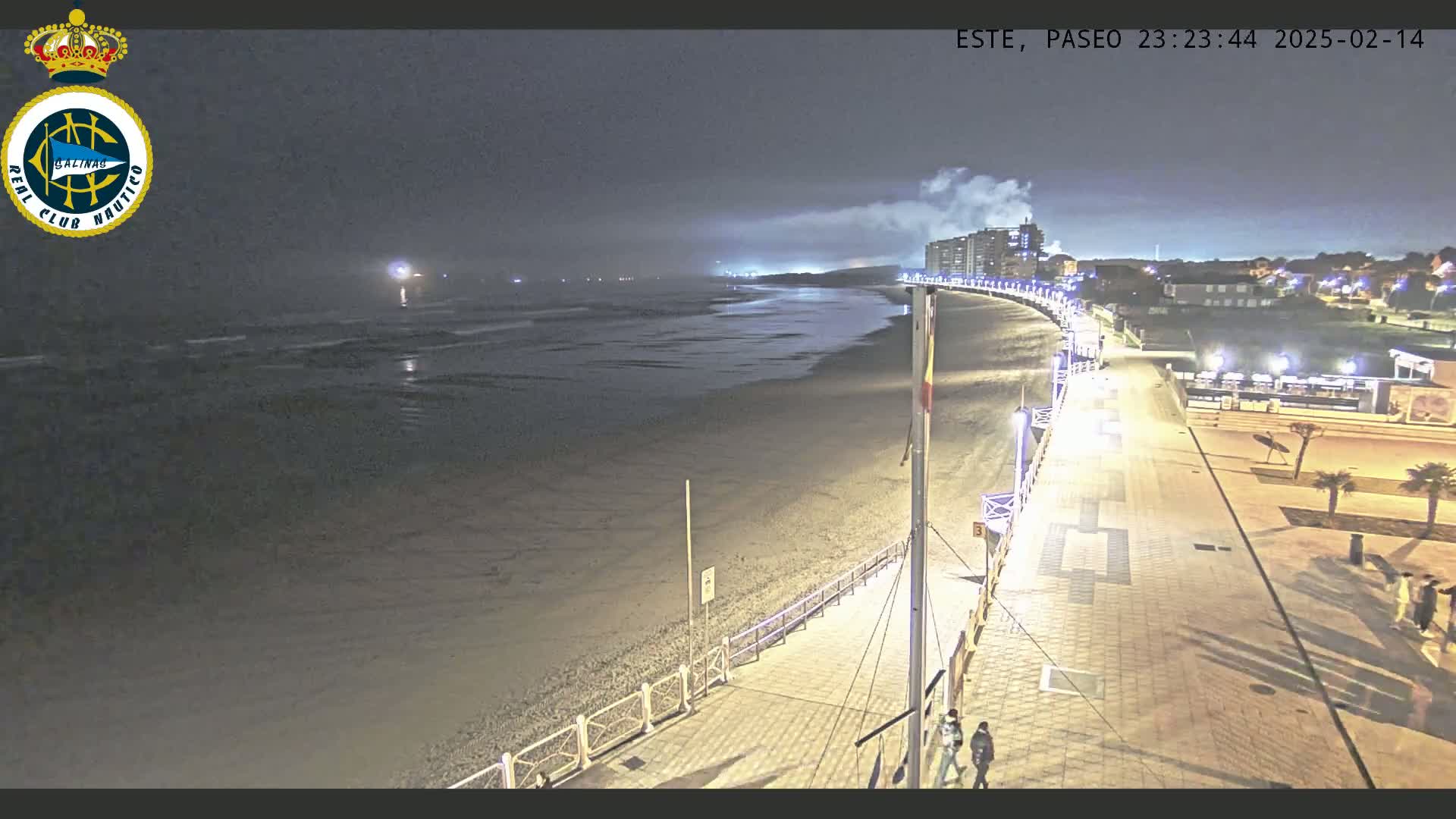 A nighttime view of a mostly empty beach and boardwalk, with hazy or foggy conditions and lights illuminating the scene.