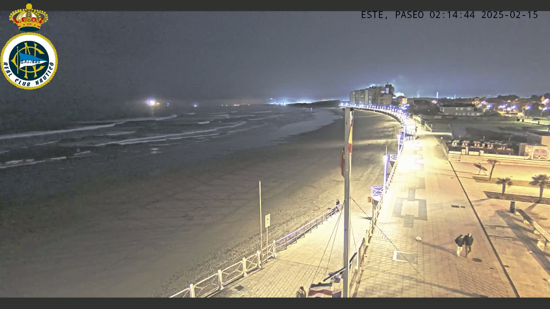 A nighttime view of a beach with gentle waves, a paved walkway lined with lights, and buildings in the distance under a dark sky.
