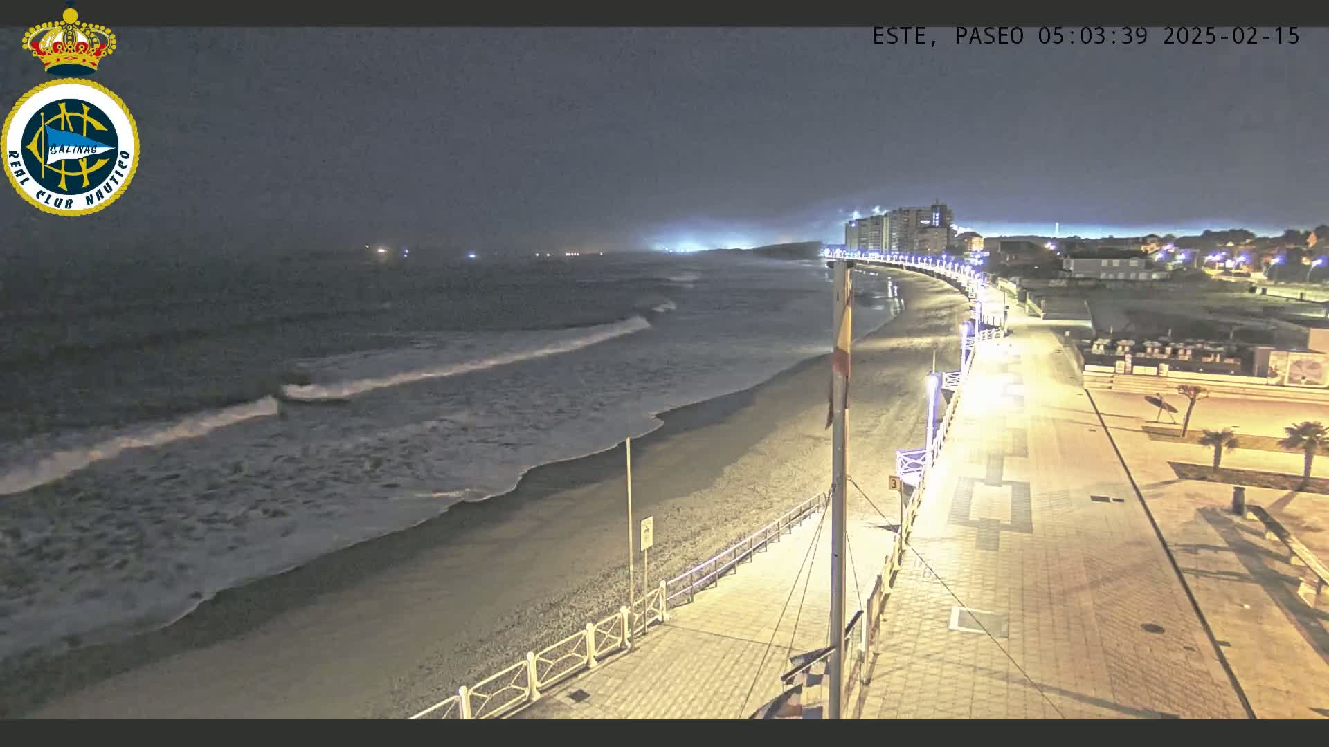 A nighttime view of a mostly empty beach with gentle waves rolling onto the shore, a paved walkway lined with lights, and buildings in the distance under a dark sky.