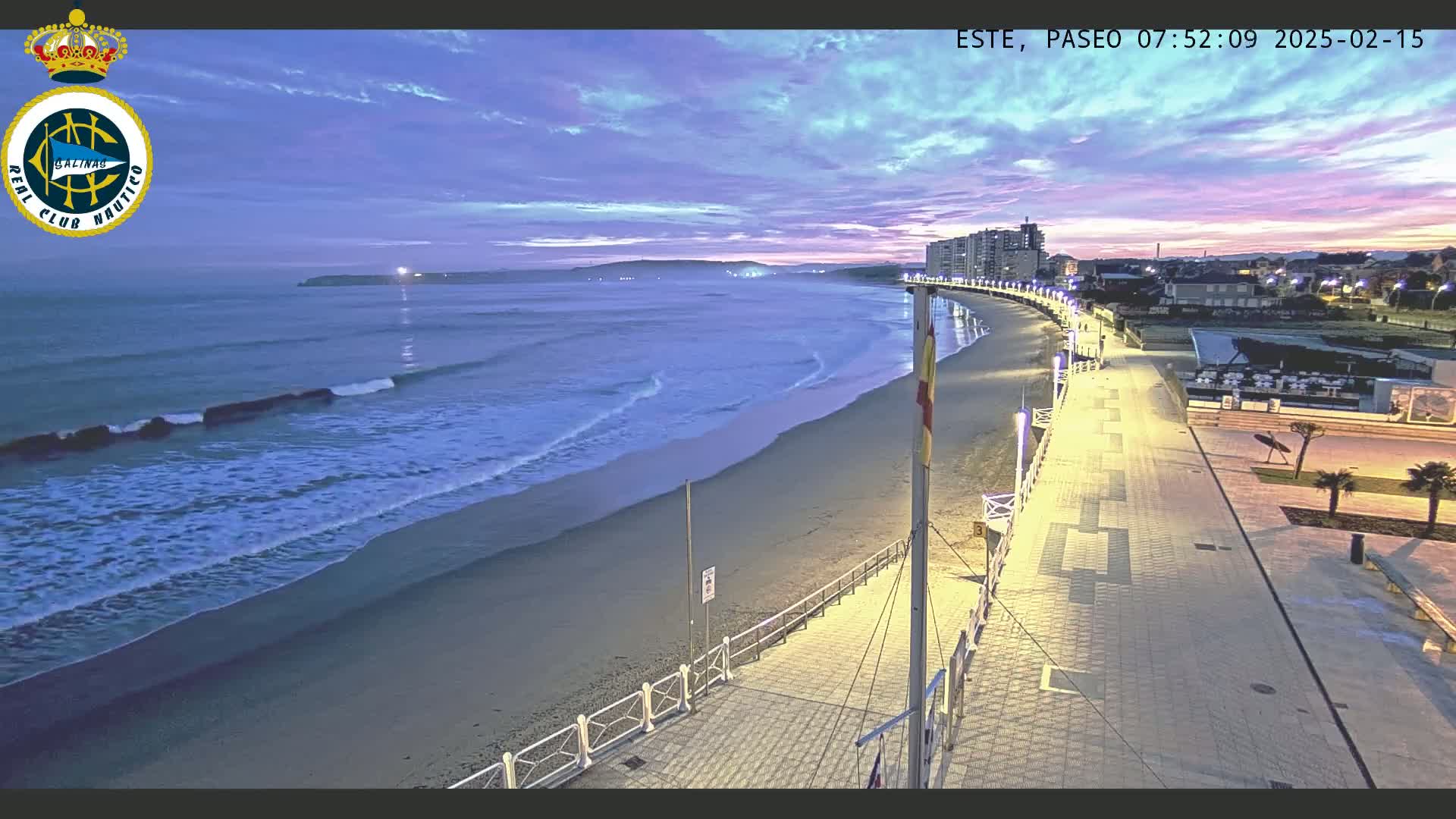 A long, sandy beach with gentle waves is illuminated at twilight by streetlights lining a paved boardwalk that leads to buildings in the distance.