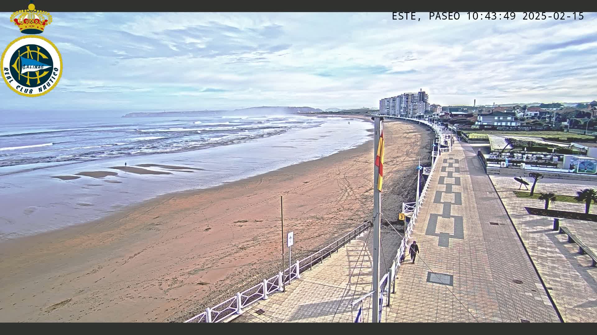 A sandy beach with gentle waves under a partly cloudy sky is bordered by a paved promenade and buildings.
