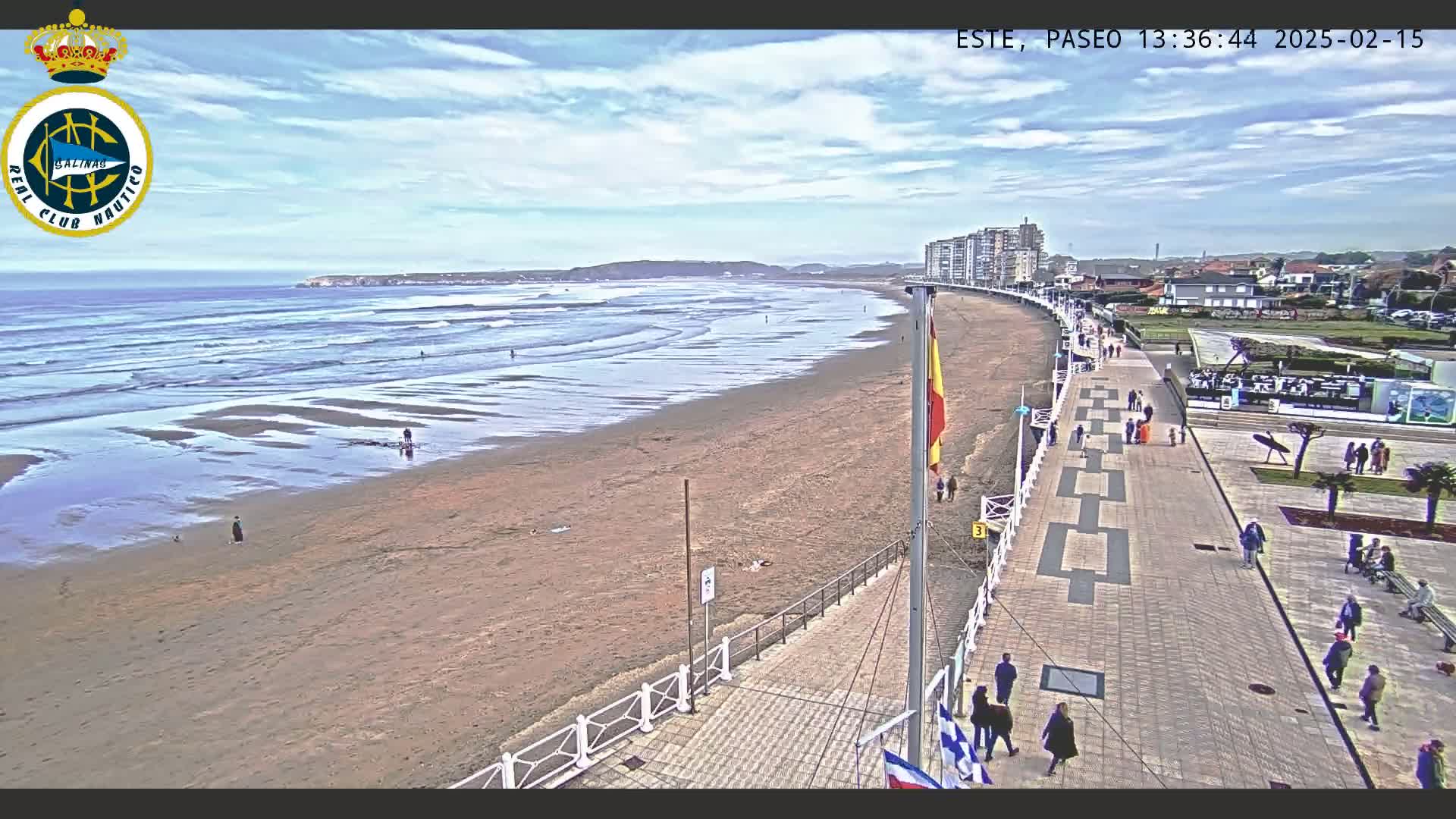 A sandy beach with small waves and a few people on it is seen from above, next to a paved walkway with people strolling along it, under a partly cloudy sky.