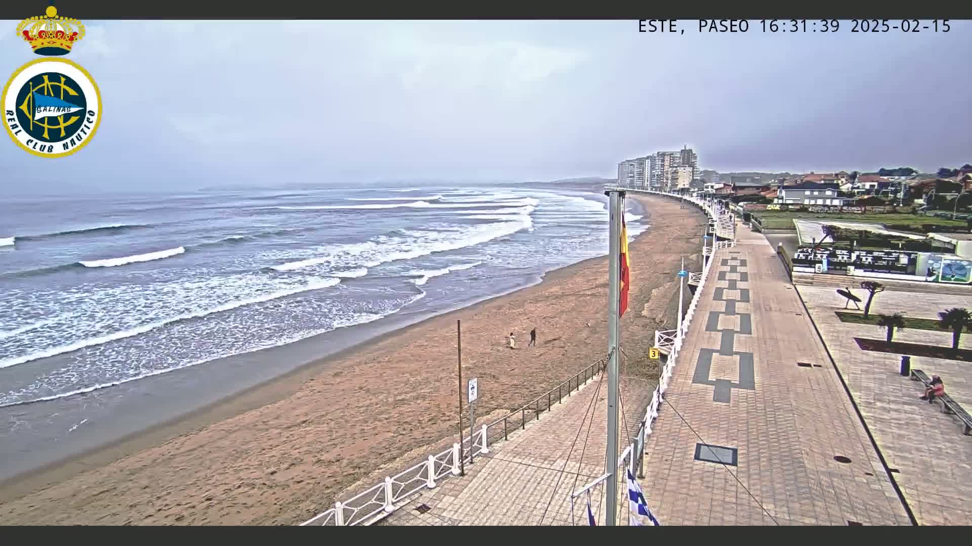 A sandy beach with moderate waves under an overcast sky is bordered by a paved walkway and buildings.
