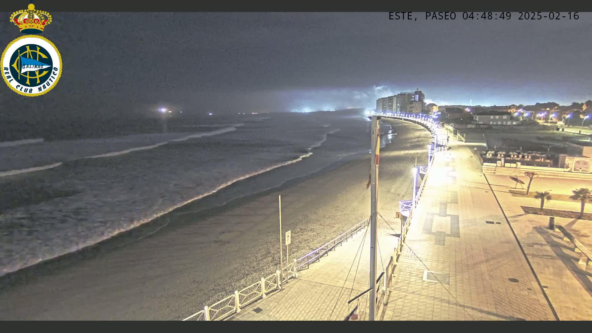 A nighttime view of a beach with gently rolling waves, a paved walkway lined with lights, and buildings in the distance under a dark, clear sky.