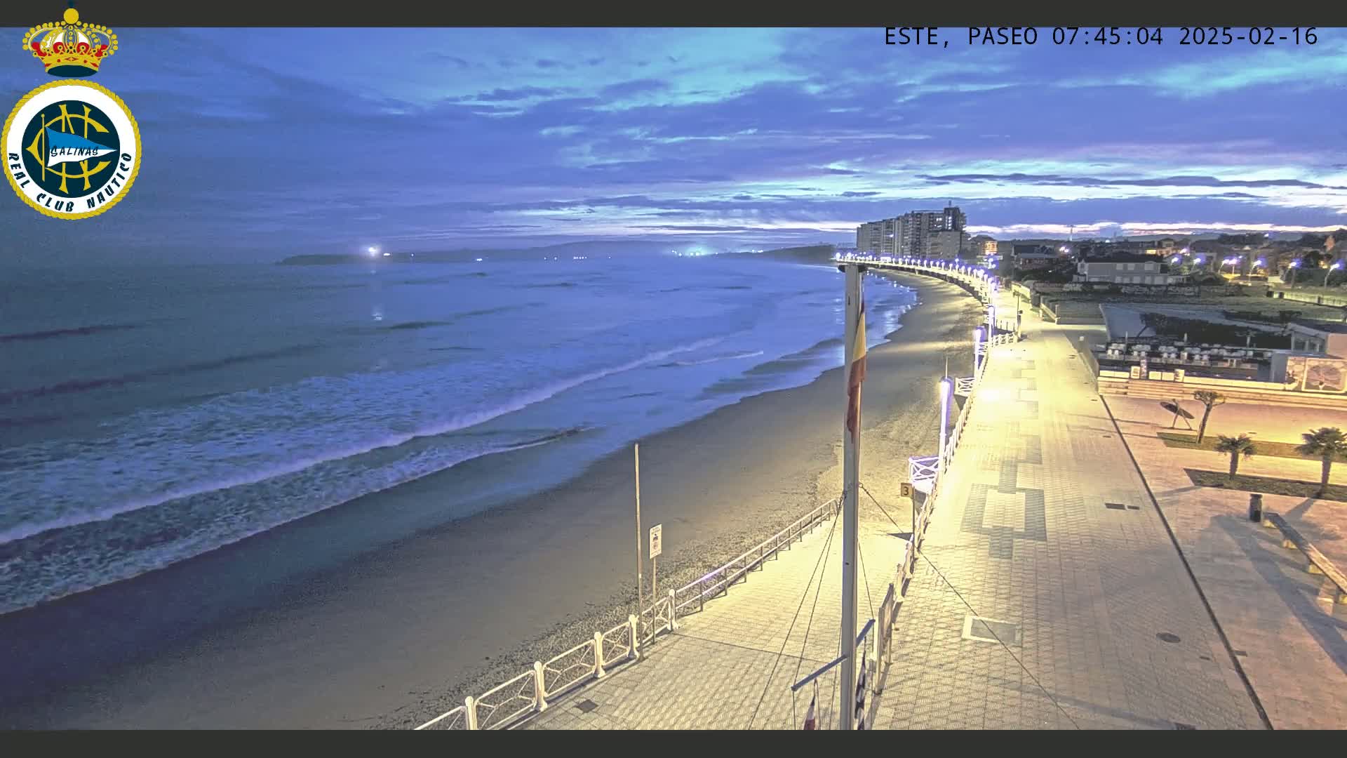 A nighttime beach scene shows gentle waves lapping the shore under a twilight sky, with a well-lit boardwalk and buildings visible in the background.