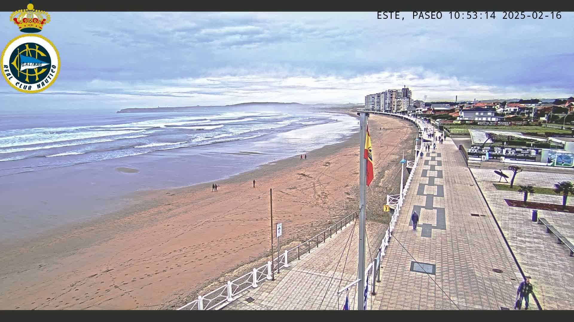 A sandy beach with waves rolling in under a partly cloudy sky is next to a paved boardwalk lined with buildings and a few pedestrians.