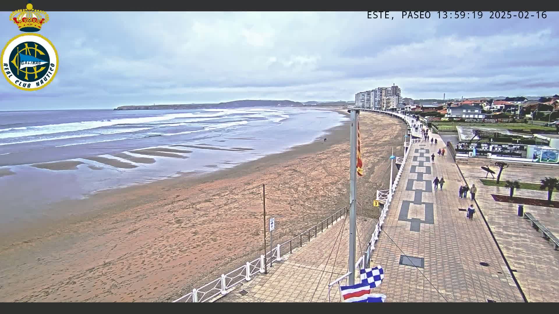 A sandy beach with gentle waves under a cloudy sky is bordered by a paved walkway with pedestrians, buildings, and green spaces.
