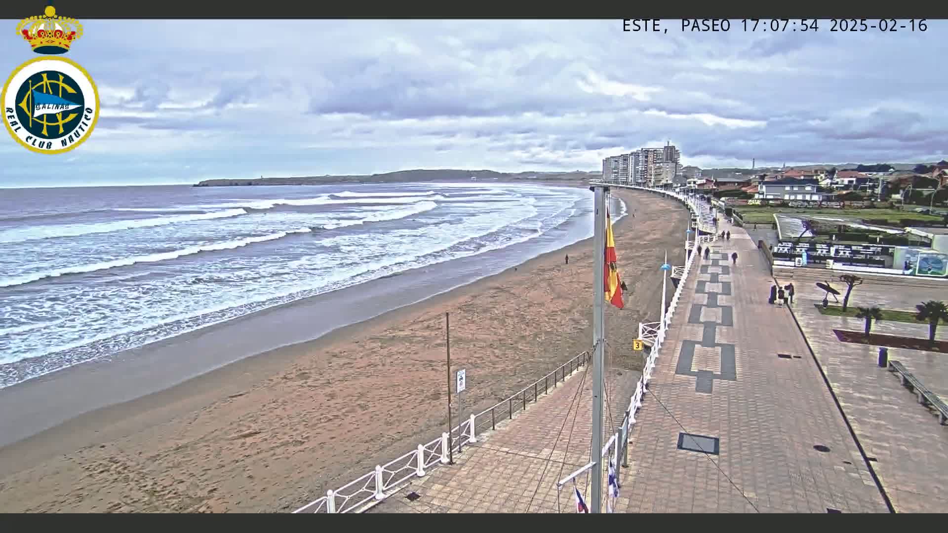 A sandy beach with moderate waves under a cloudy sky is bordered by a paved walkway with people strolling, and buildings are visible in the background.