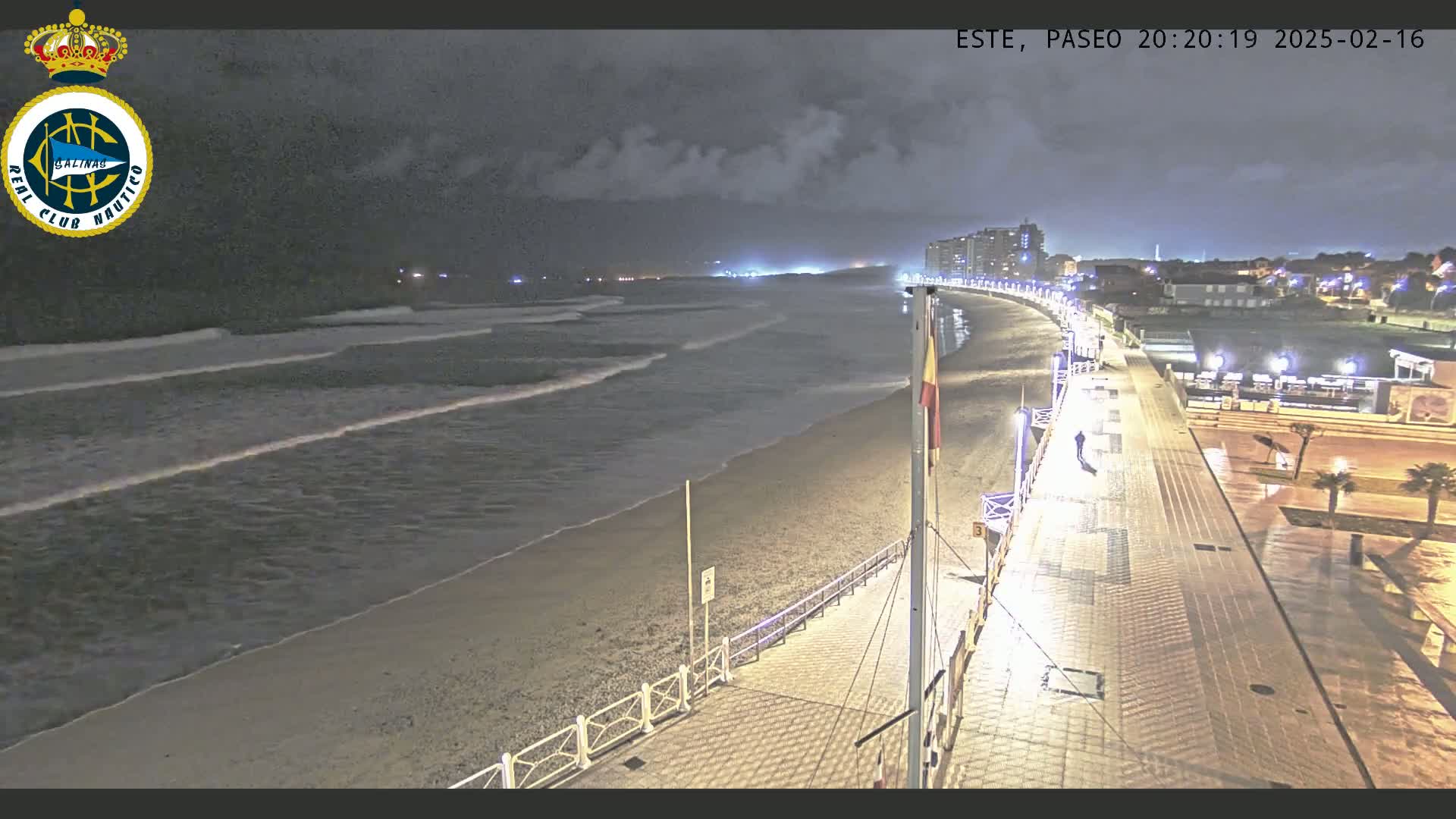 A nighttime view of a beach with gentle waves lapping the shore, a paved walkway lined with lights, and buildings in the distance under a partly cloudy sky.