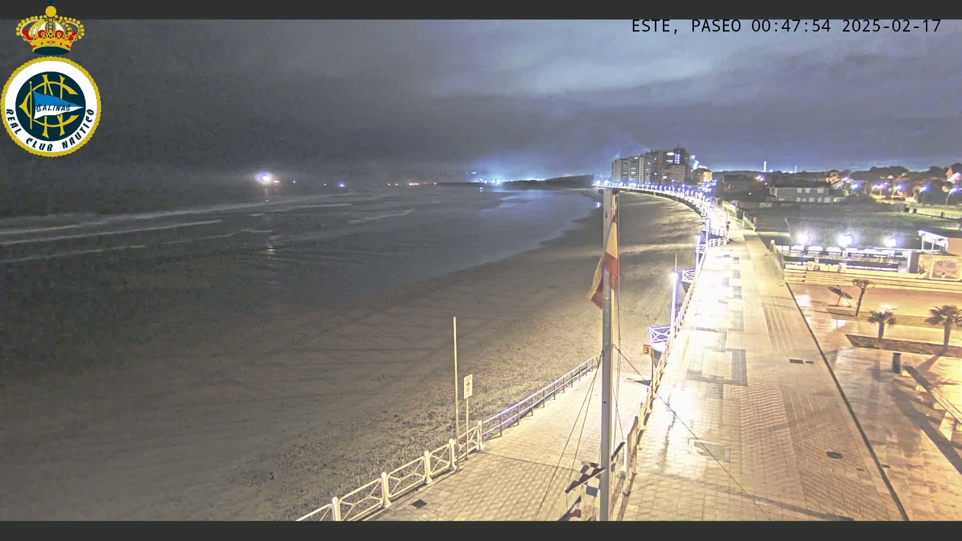 A nighttime view of a mostly empty beach with a paved walkway and buildings in the distance under a cloudy sky.