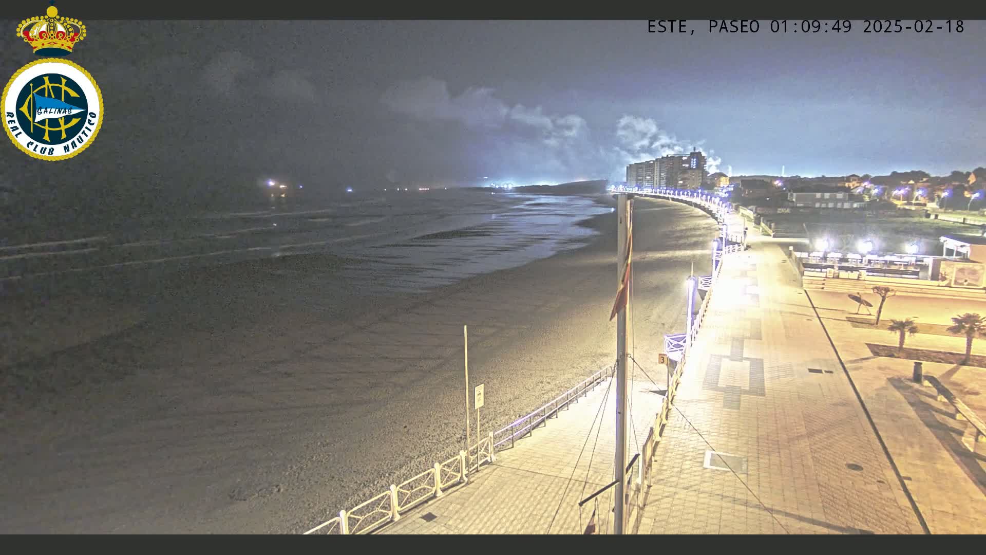 A nighttime view of a dark, sandy beach with calm waves, a boardwalk lined with lights, and buildings in the distance under a cloudy sky.