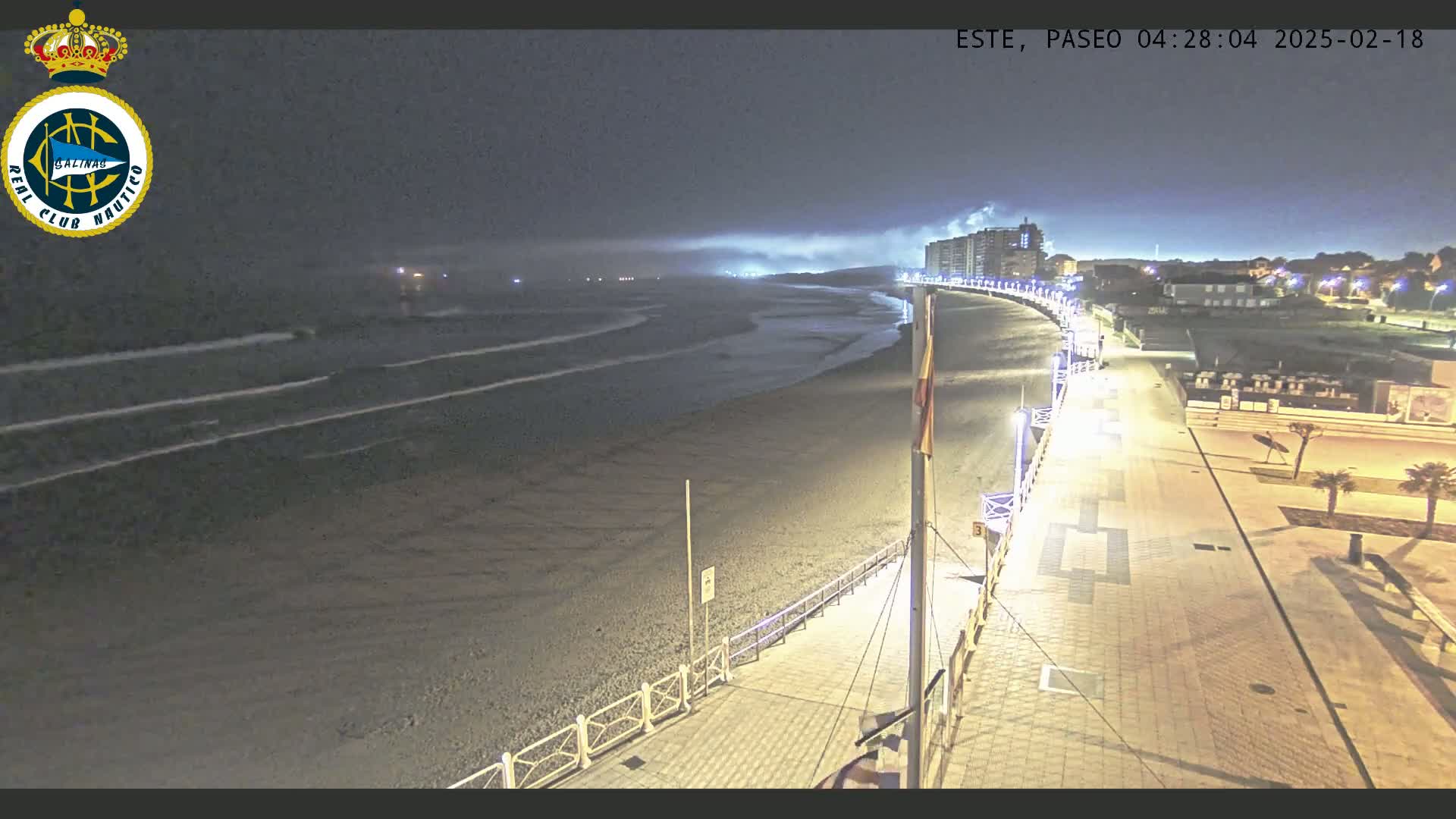 A nighttime, high-angle view shows a mostly empty beach with gentle waves, a paved walkway with lights, and buildings in the background under a dark, cloudy sky.