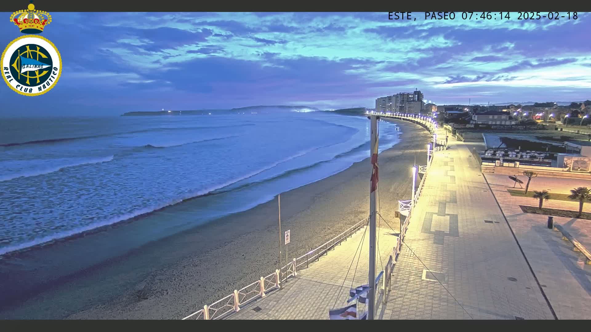 A twilight view shows a sandy beach with gentle waves lapping the shore, next to a paved promenade lined with lights, under a mostly clear sky.