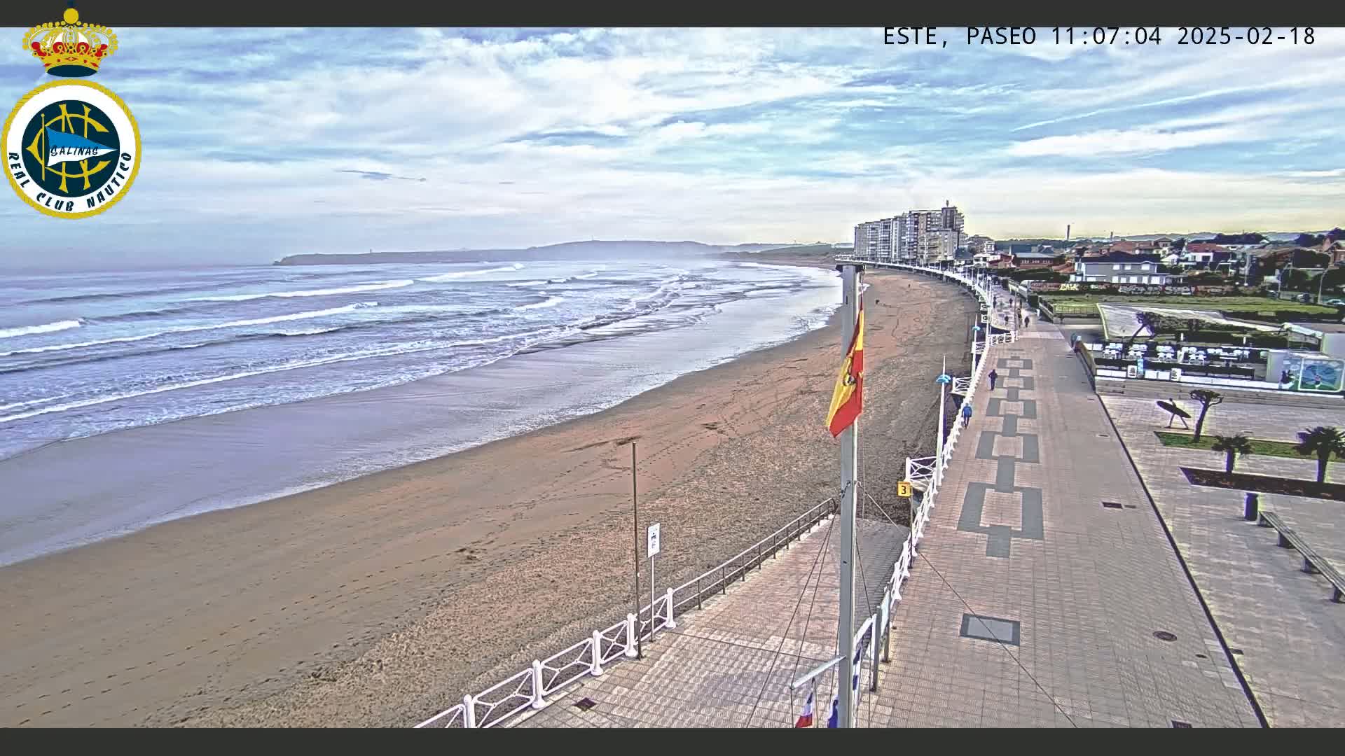 A sandy beach with moderate waves under a partly cloudy sky is next to a paved boardwalk lined with buildings, with a Spanish flag waving in the breeze.