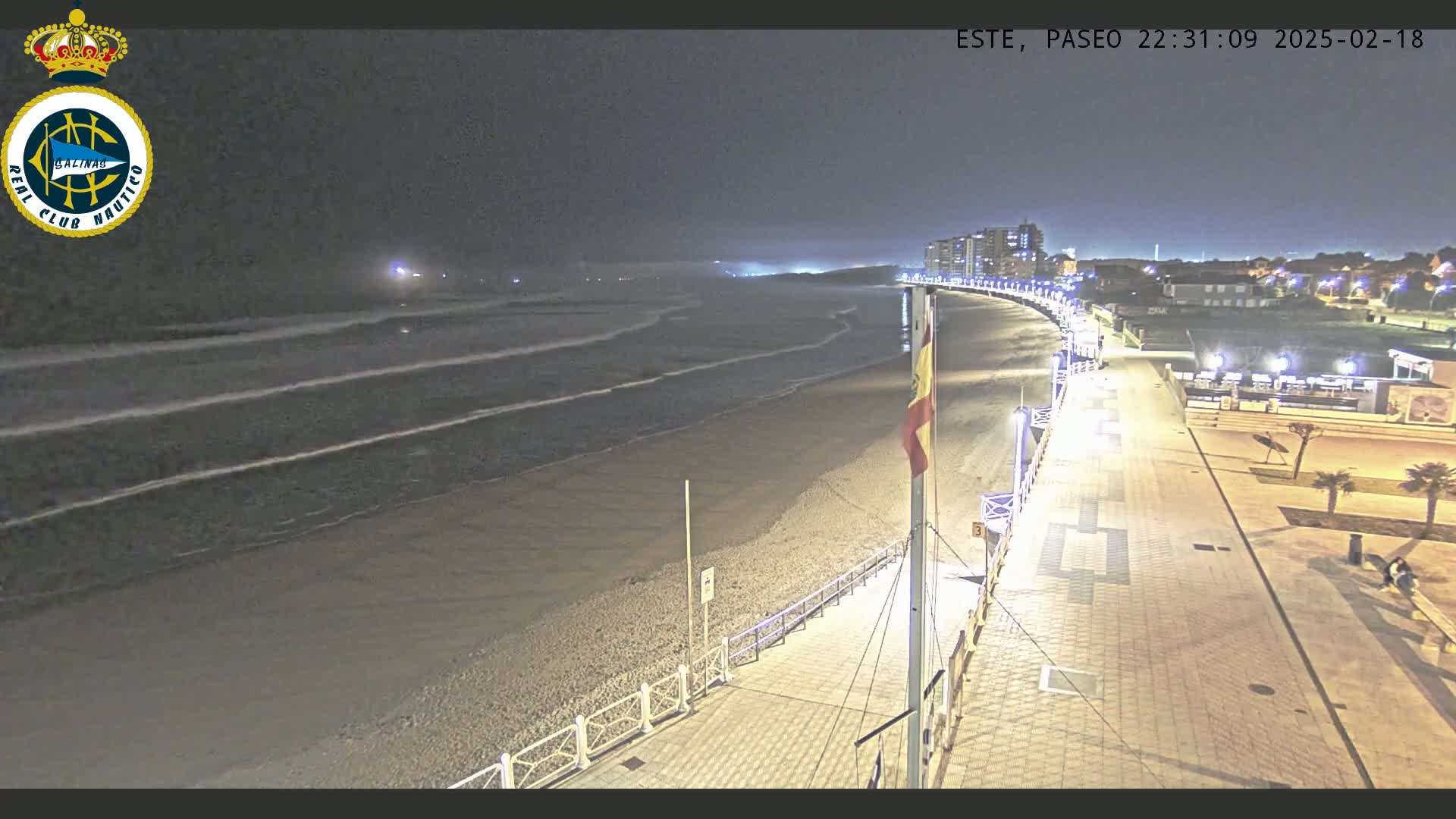 A nighttime view shows a beach with gentle waves lapping the shore, a paved walkway with lights lining it, and buildings in the distance.