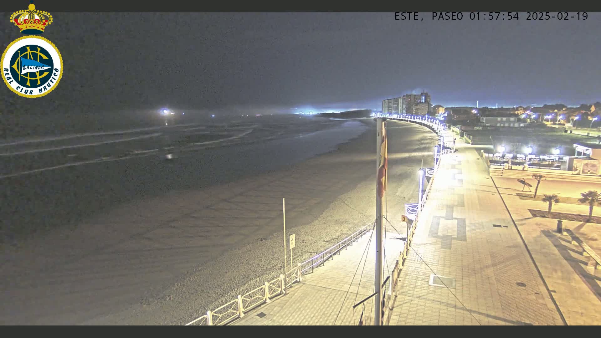 A nighttime view of a beach with a paved walkway and buildings in the distance under a dark, cloudy sky.