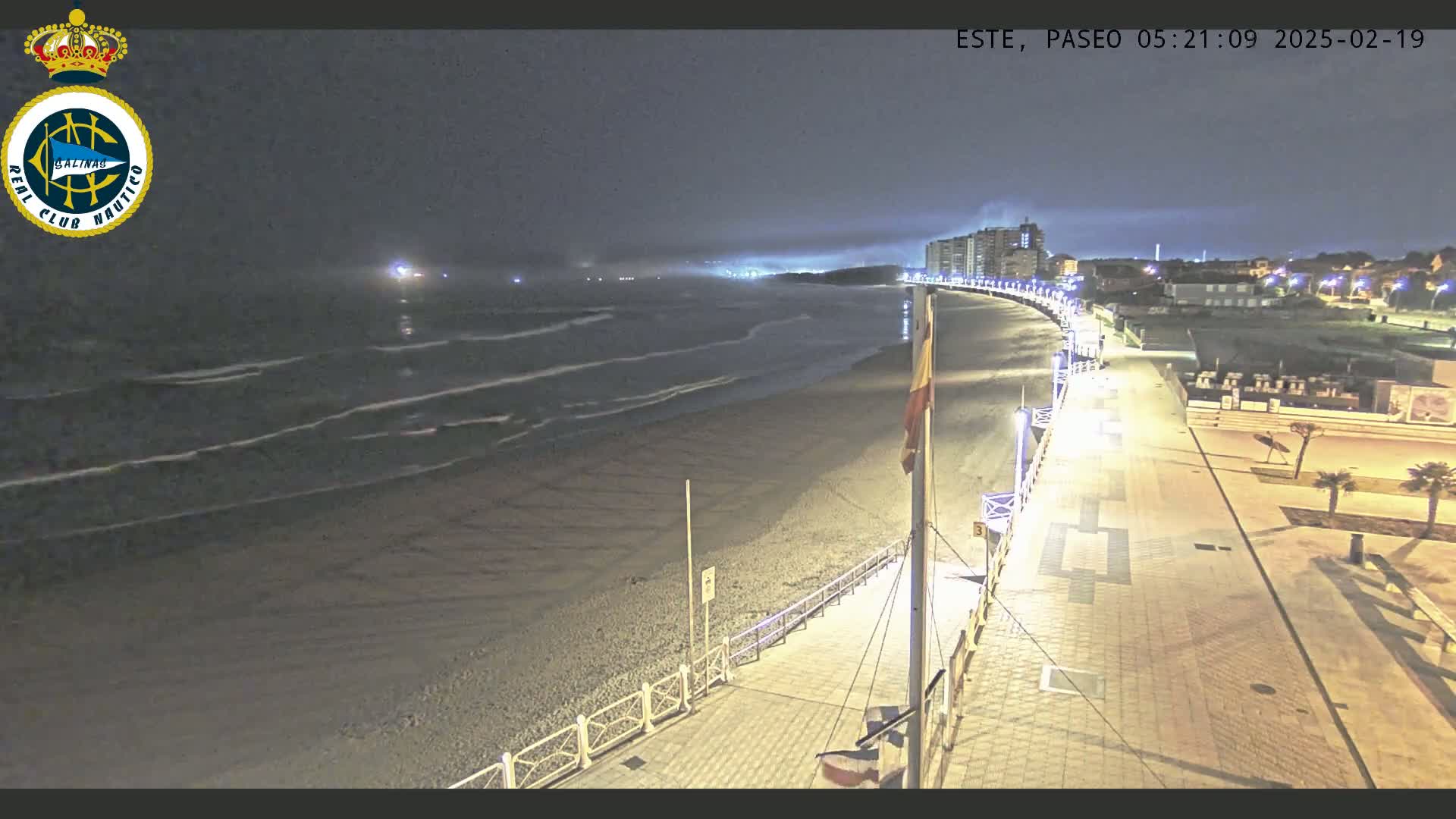 A nighttime view of a beach at low tide with gentle waves, a brightly lit boardwalk, and buildings in the distance under a dark, cloudy sky.