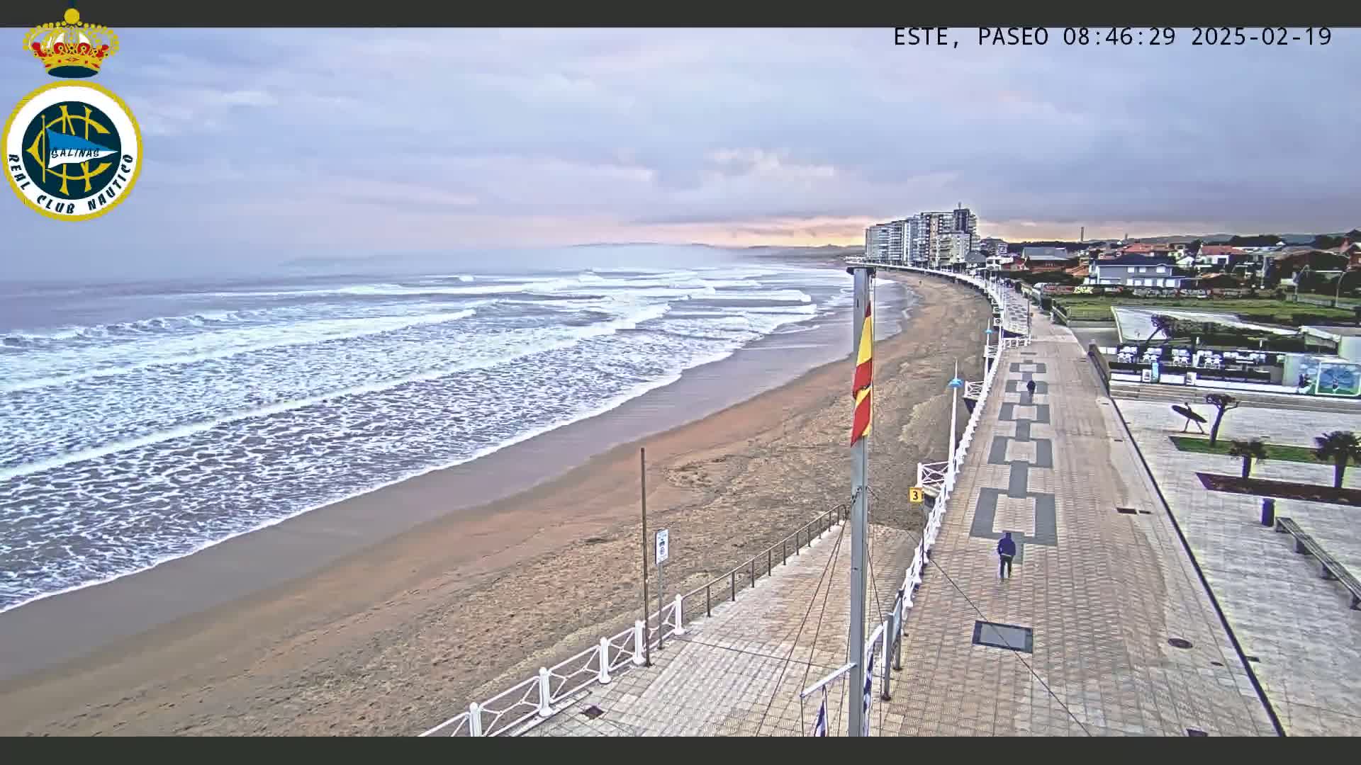 A sandy beach with moderate waves under an overcast sky is bordered by a paved walkway and buildings.