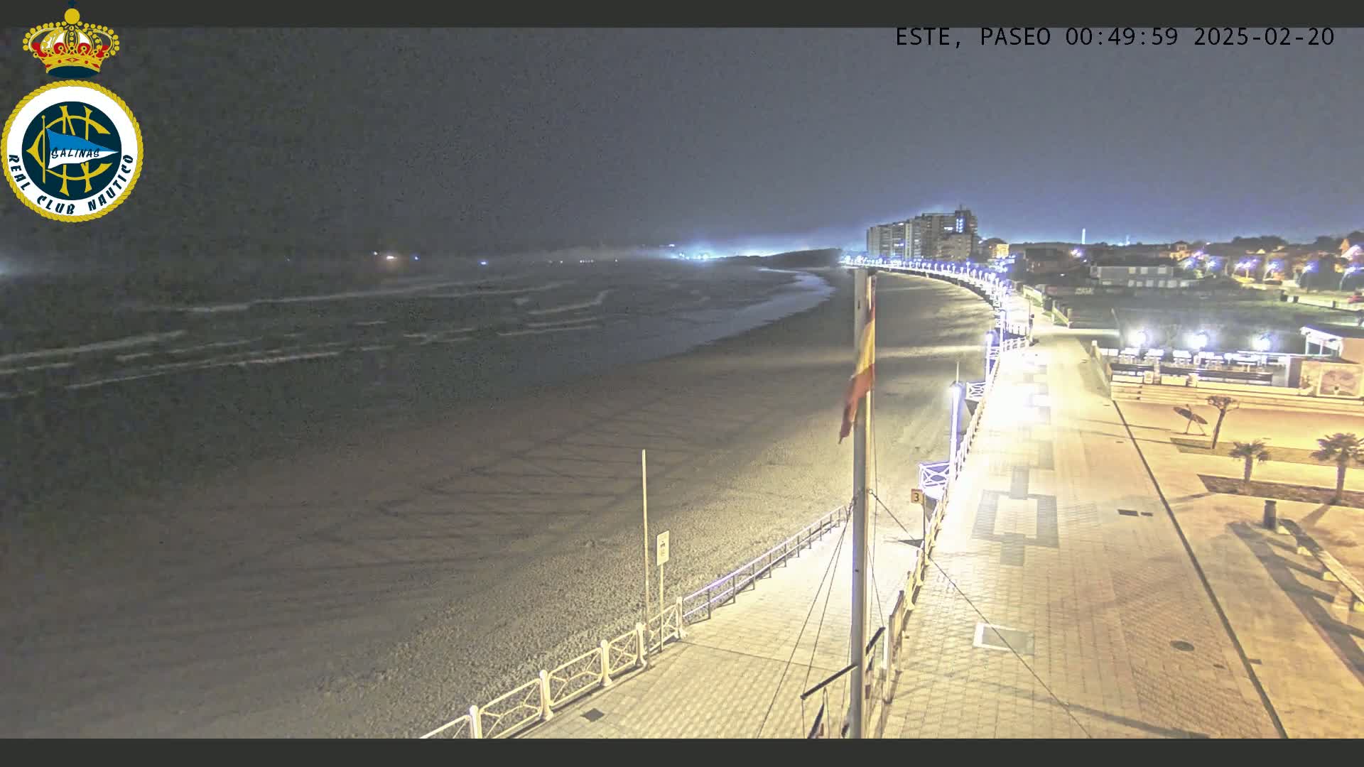 A nighttime view of a mostly empty beach with a paved walkway and buildings in the distance under a dark, cloudy sky.