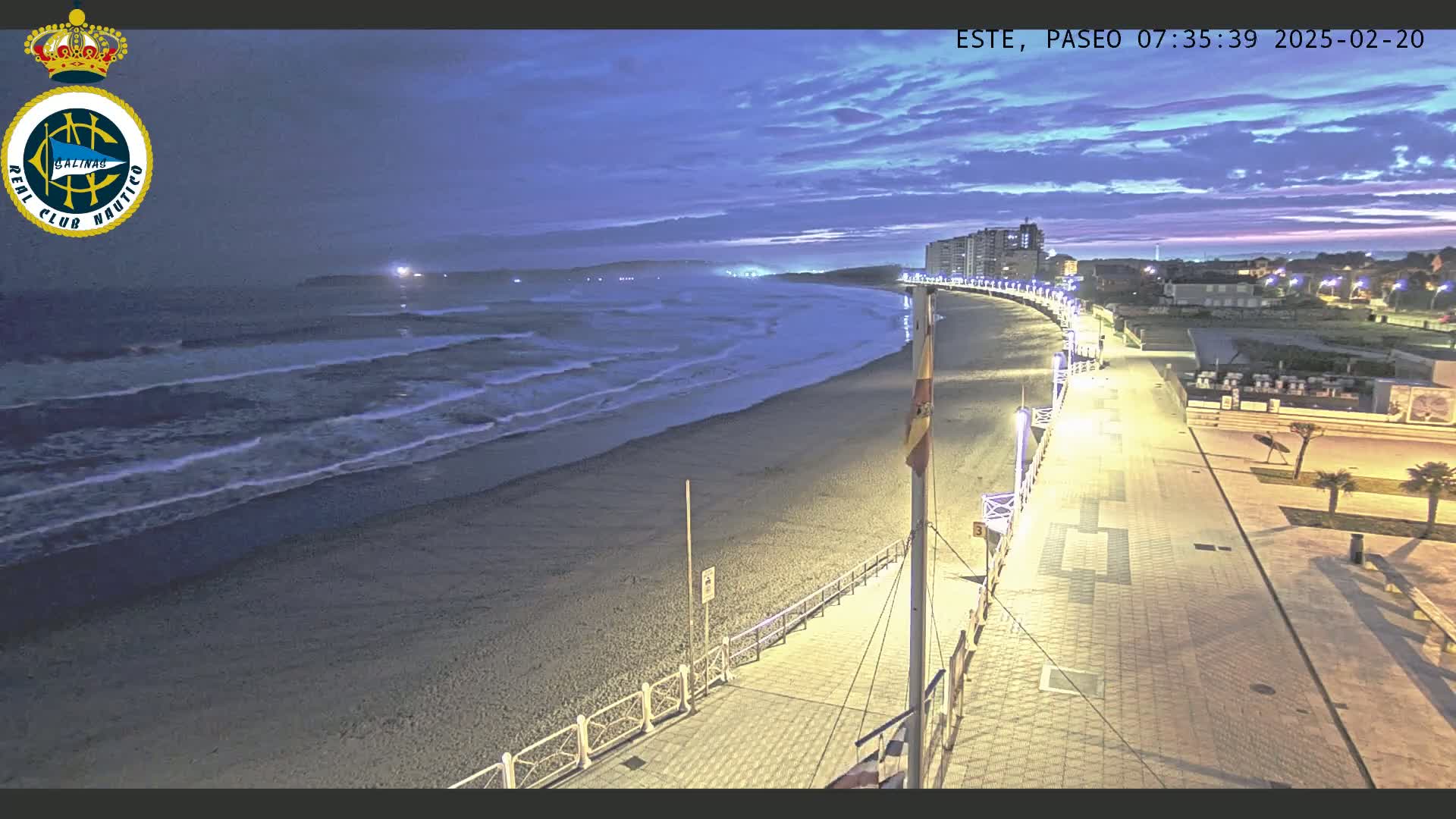 A mostly sandy beach at twilight with gentle waves rolling in, a paved boardwalk lit at night, and buildings in the background.