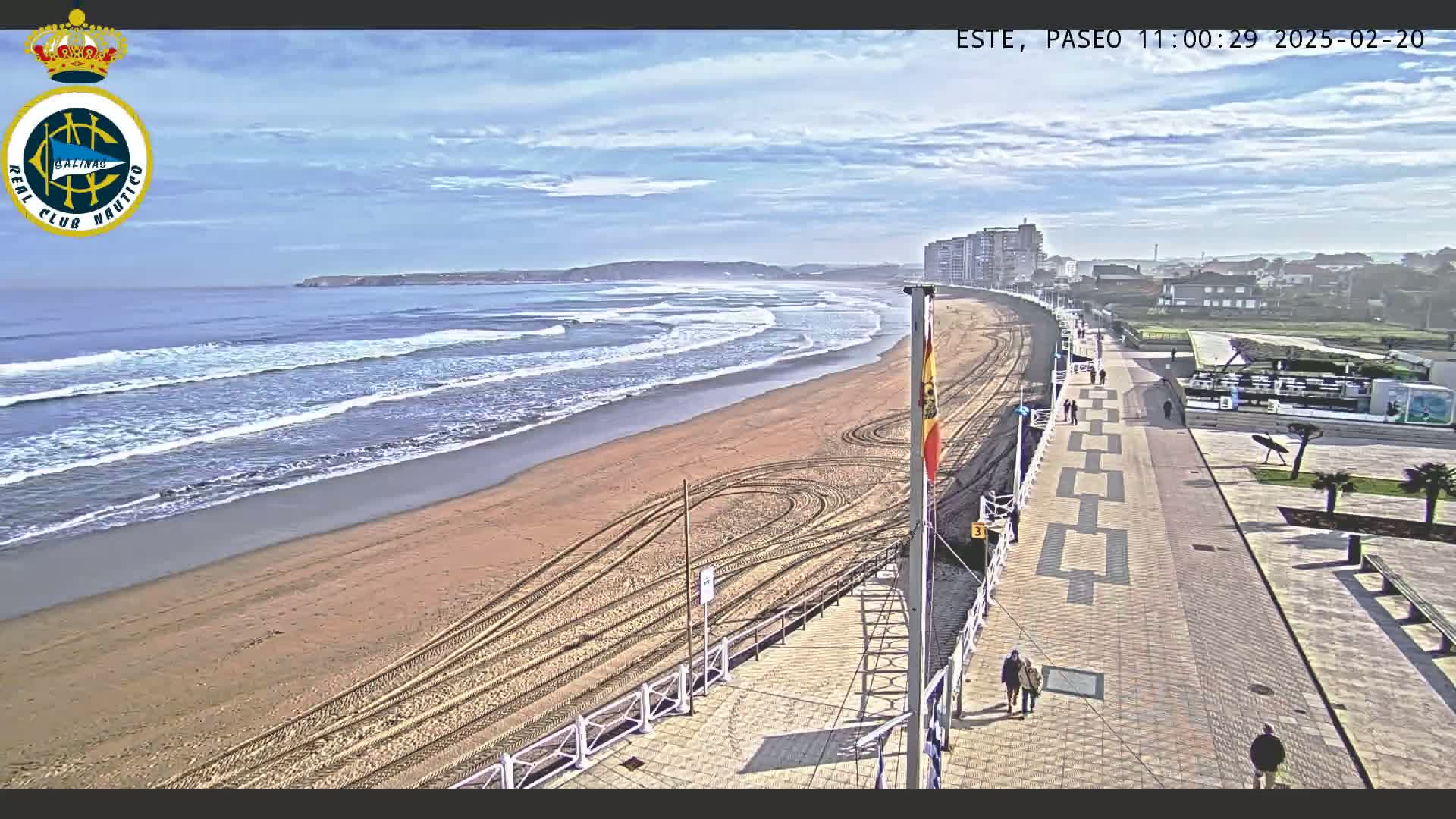 A sandy beach with gentle waves under a mostly clear sky is bordered by a paved walkway with several people walking, and buildings are visible in the distance.