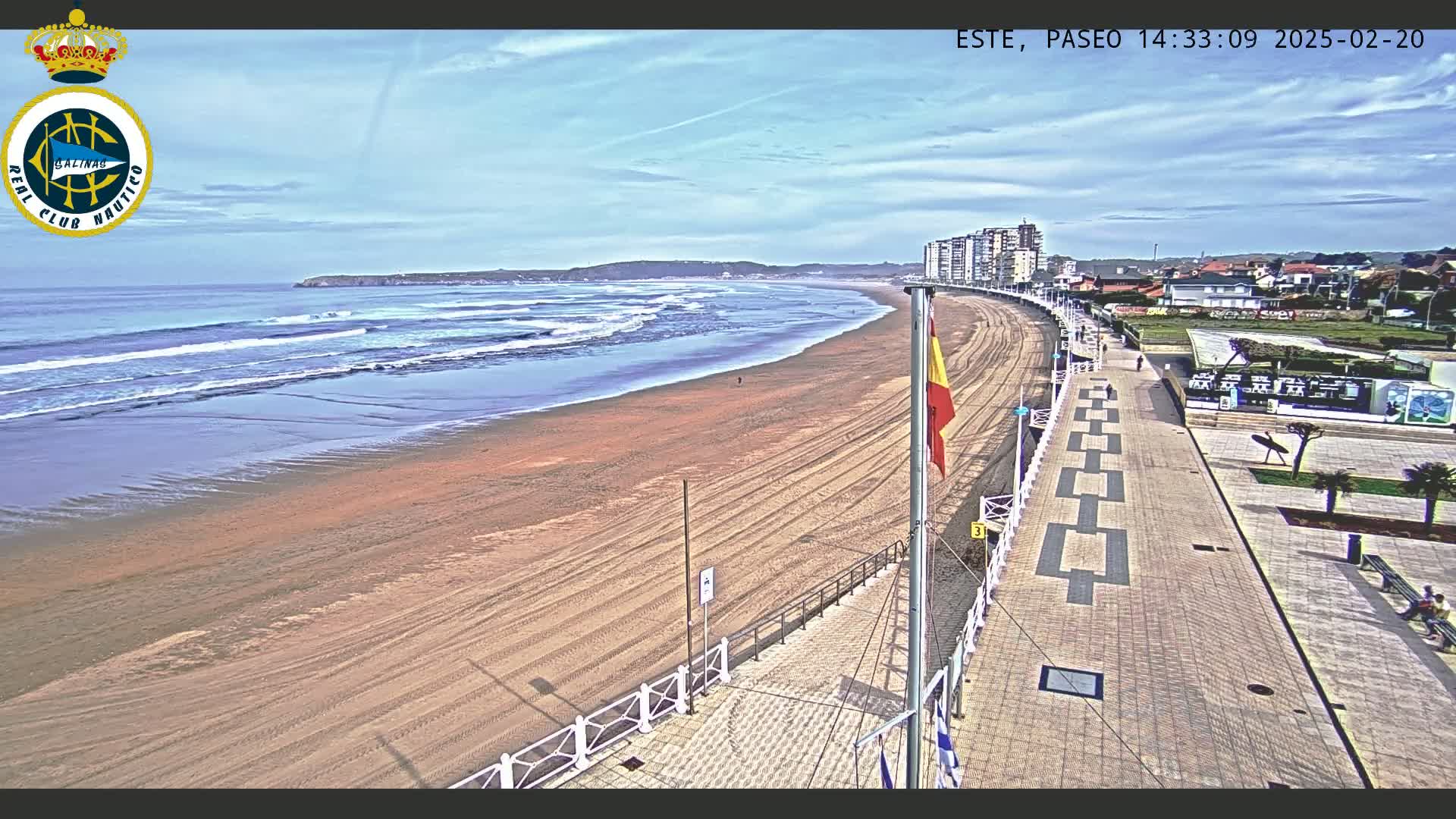 A sandy beach with gentle waves under a mostly clear sky is bordered by a paved boardwalk and buildings.