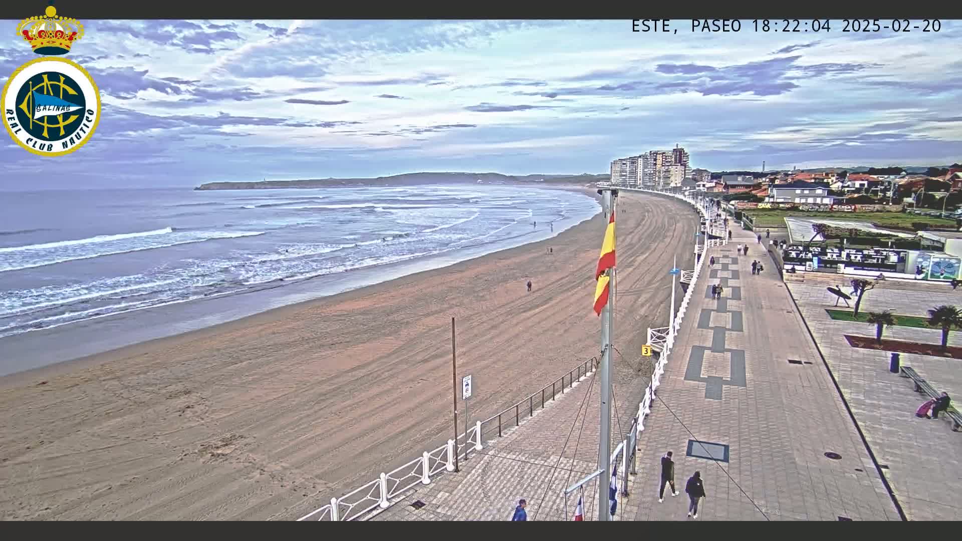 A sandy beach with gentle waves under a partly cloudy sky is bordered by a paved promenade with a few pedestrians, and buildings are visible in the background.