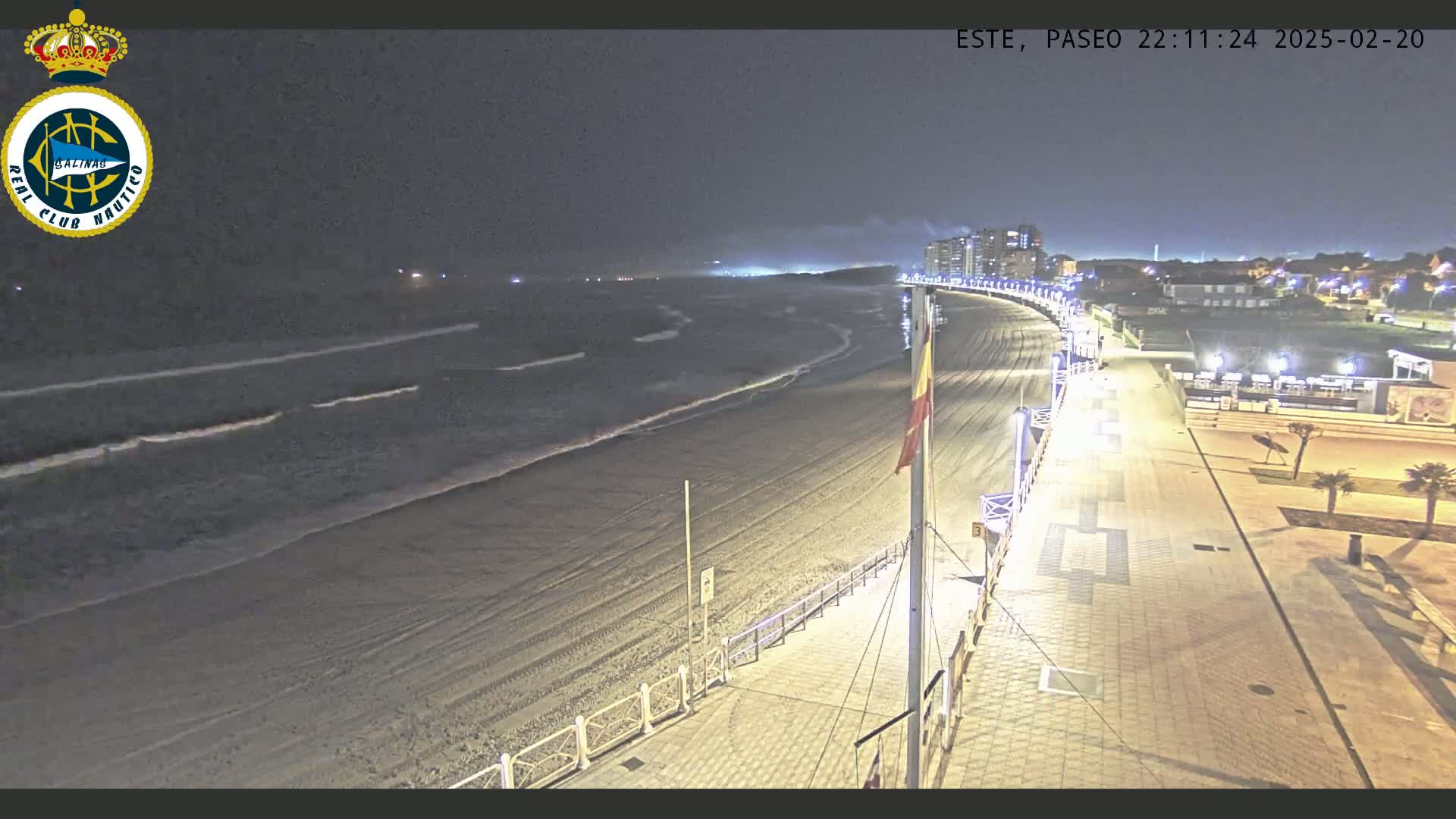 A nighttime view of a sandy beach with gentle waves, a paved boardwalk with lights, and buildings in the distance under a dark sky.