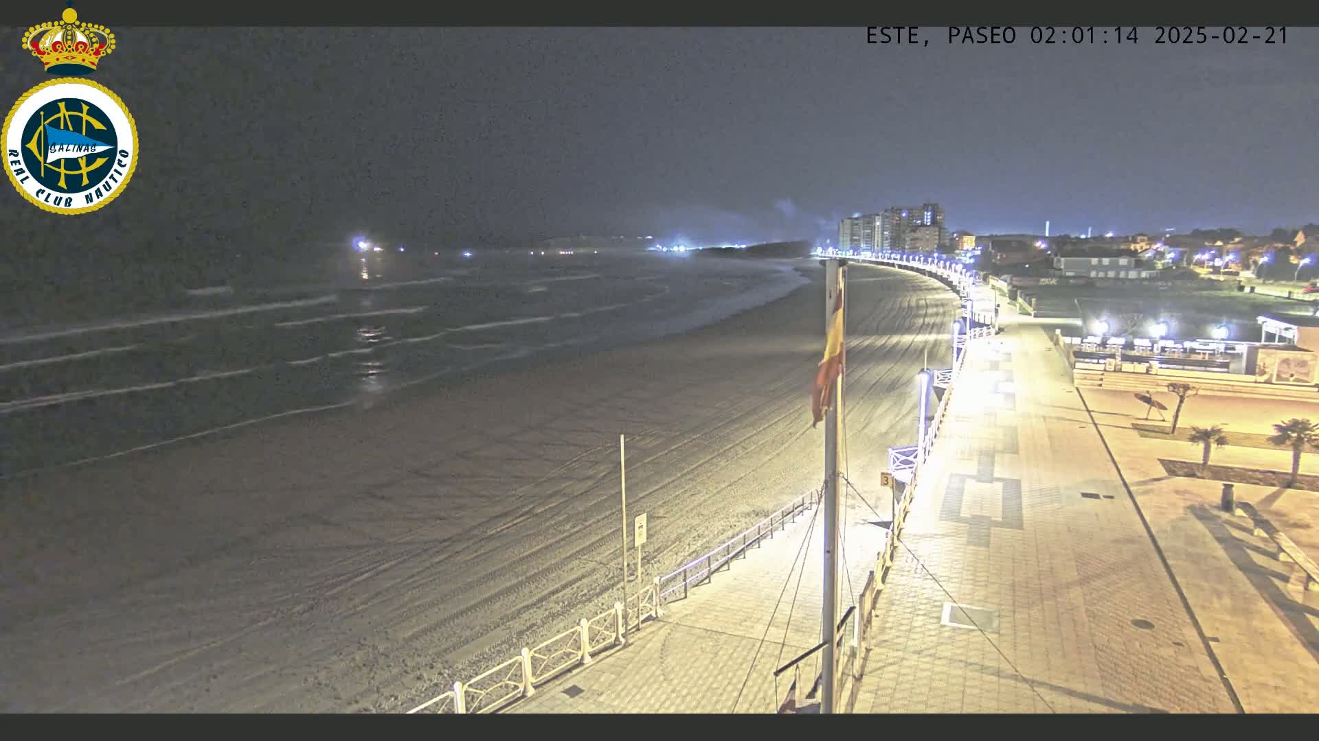 A nighttime view of a sandy beach with gentle waves, a paved boardwalk, and buildings in the distance under a dark sky.
