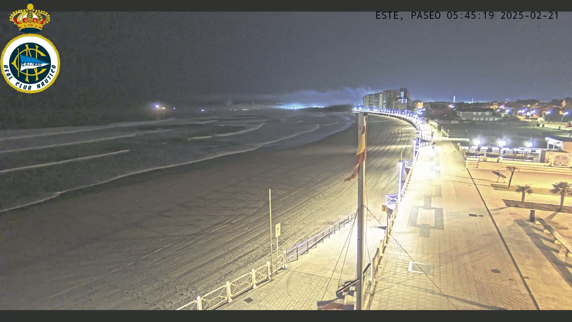 A nighttime view of a beach with gently lapping waves and a paved walkway lit by streetlights, under a slightly hazy sky.