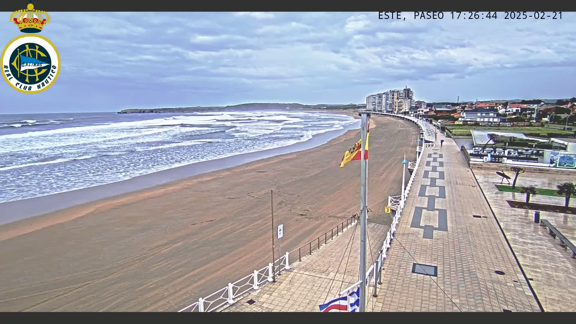 A sandy beach with moderate waves under a partly cloudy sky is seen from an elevated vantage point overlooking a paved boardwalk and buildings.