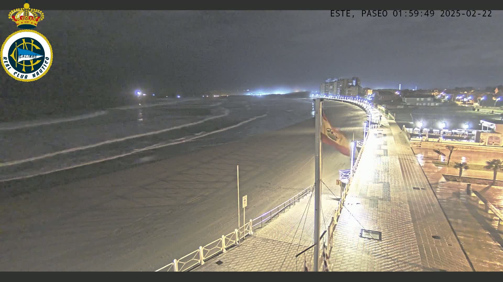 A nighttime view of a wet beach and boardwalk, with gentle waves lapping the shore and buildings visible in the distance under a dark sky.