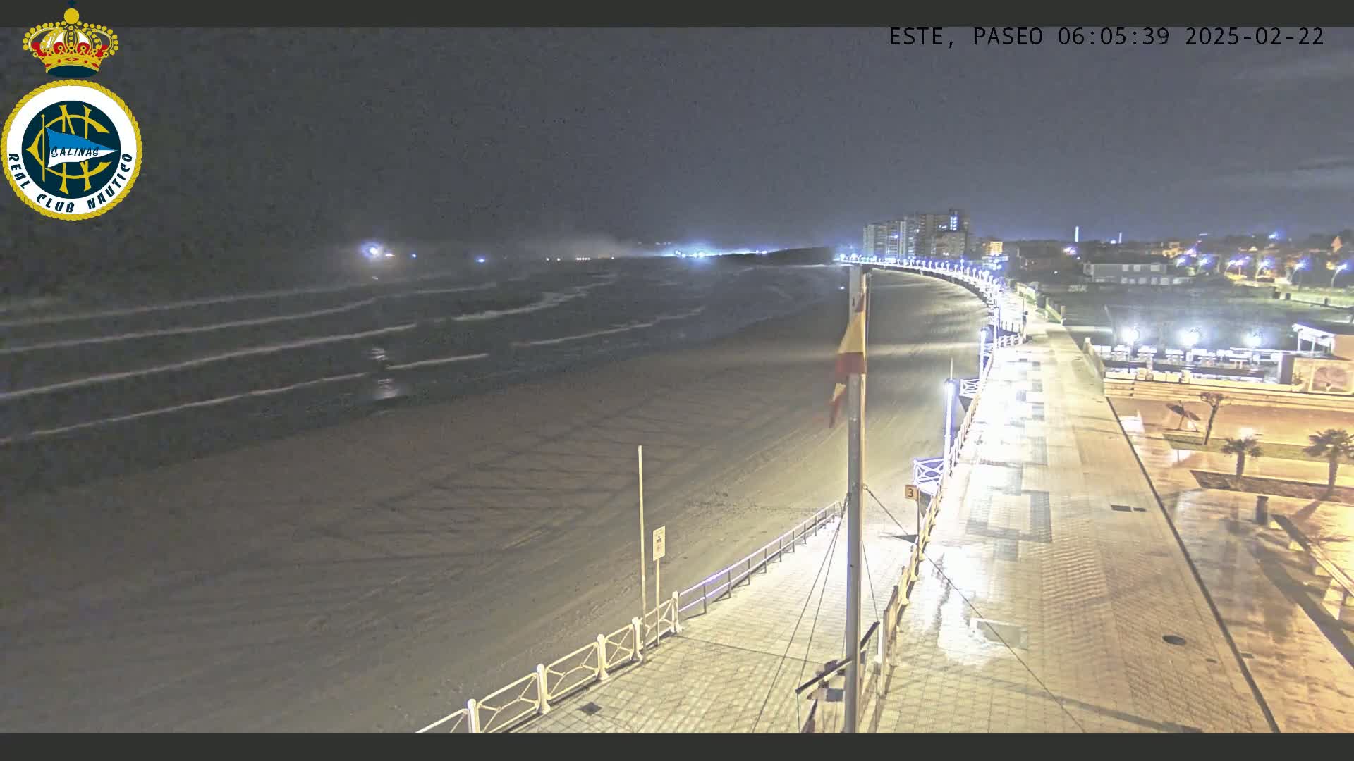 A nighttime, high-angle view shows a mostly empty beach with moderate waves under a dark sky, next to a well-lit paved boardwalk that curves towards buildings in the distance.