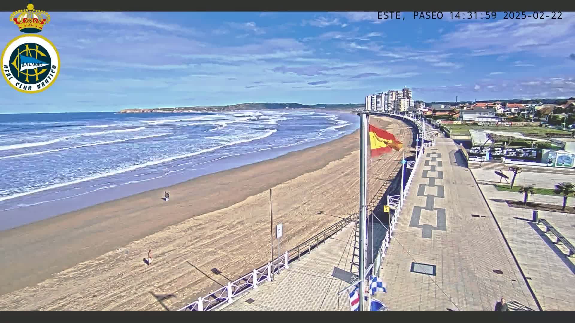 A sandy beach with gentle waves under a sunny sky is adjacent to a paved boardwalk lined with buildings and a Spanish flag.