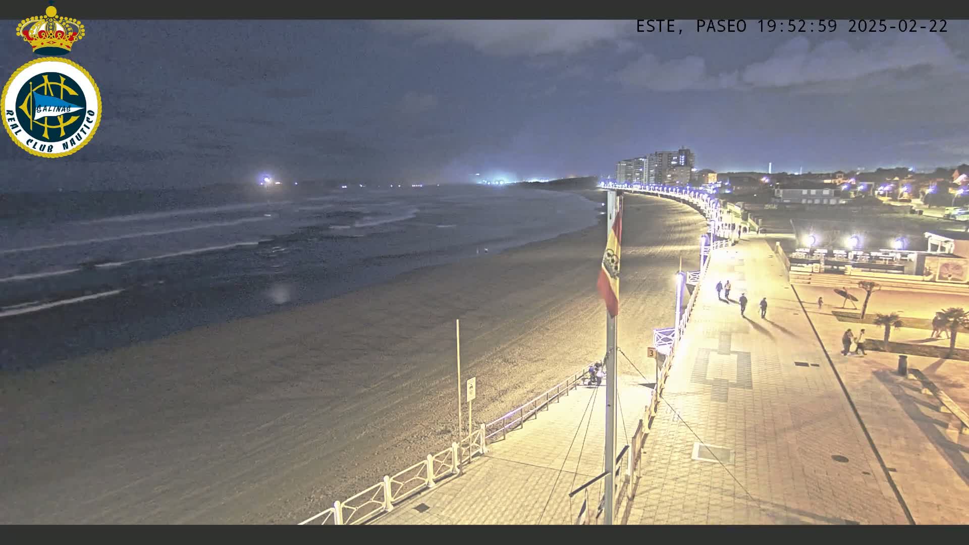 A nighttime view of a beach with moderate waves, a paved boardwalk with several people walking, and buildings in the distance under a dark sky.