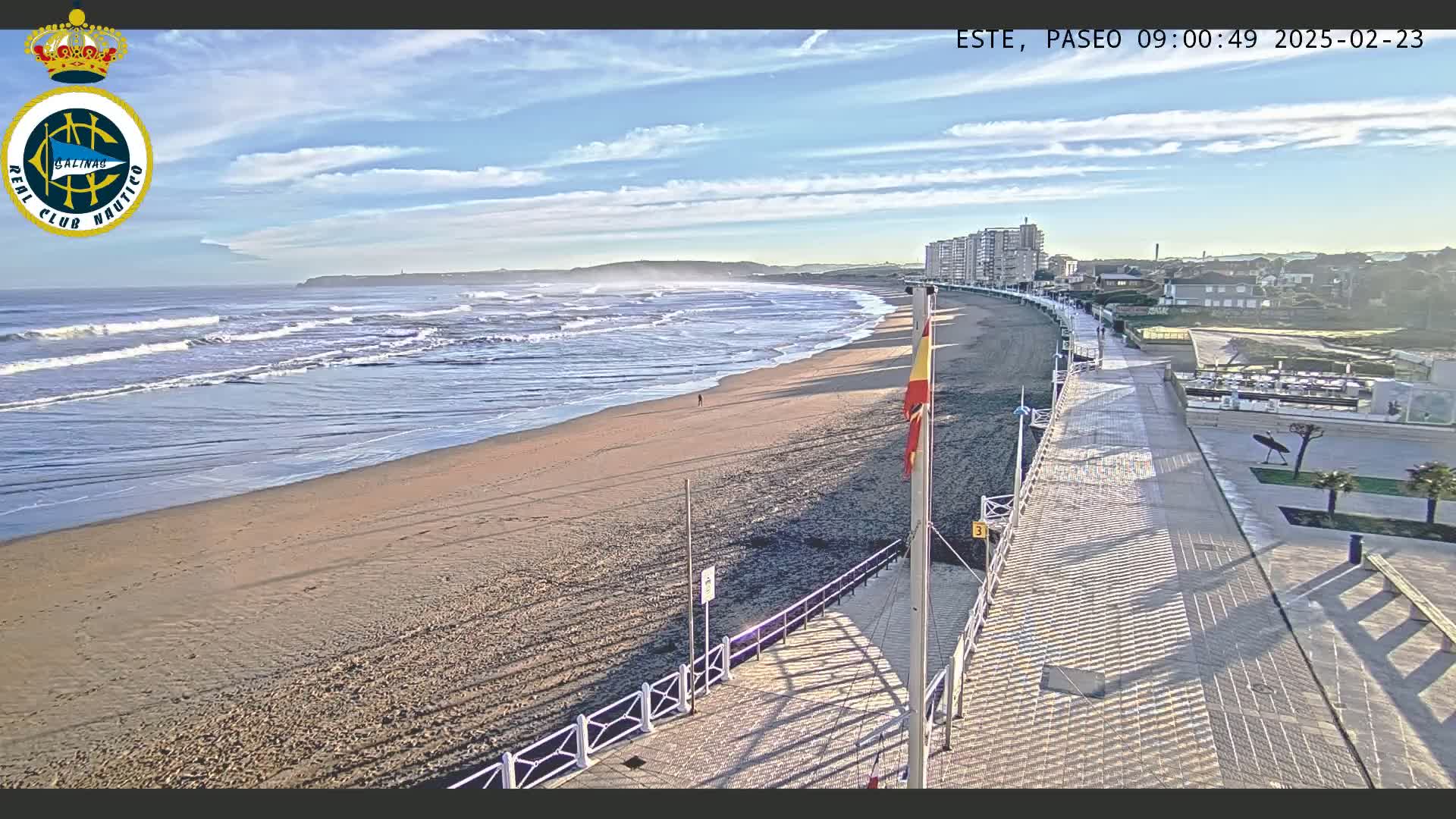 A sandy beach with moderate waves under a mostly sunny sky is bordered by a paved walkway leading to buildings, with a flagpole situated near the walkway.