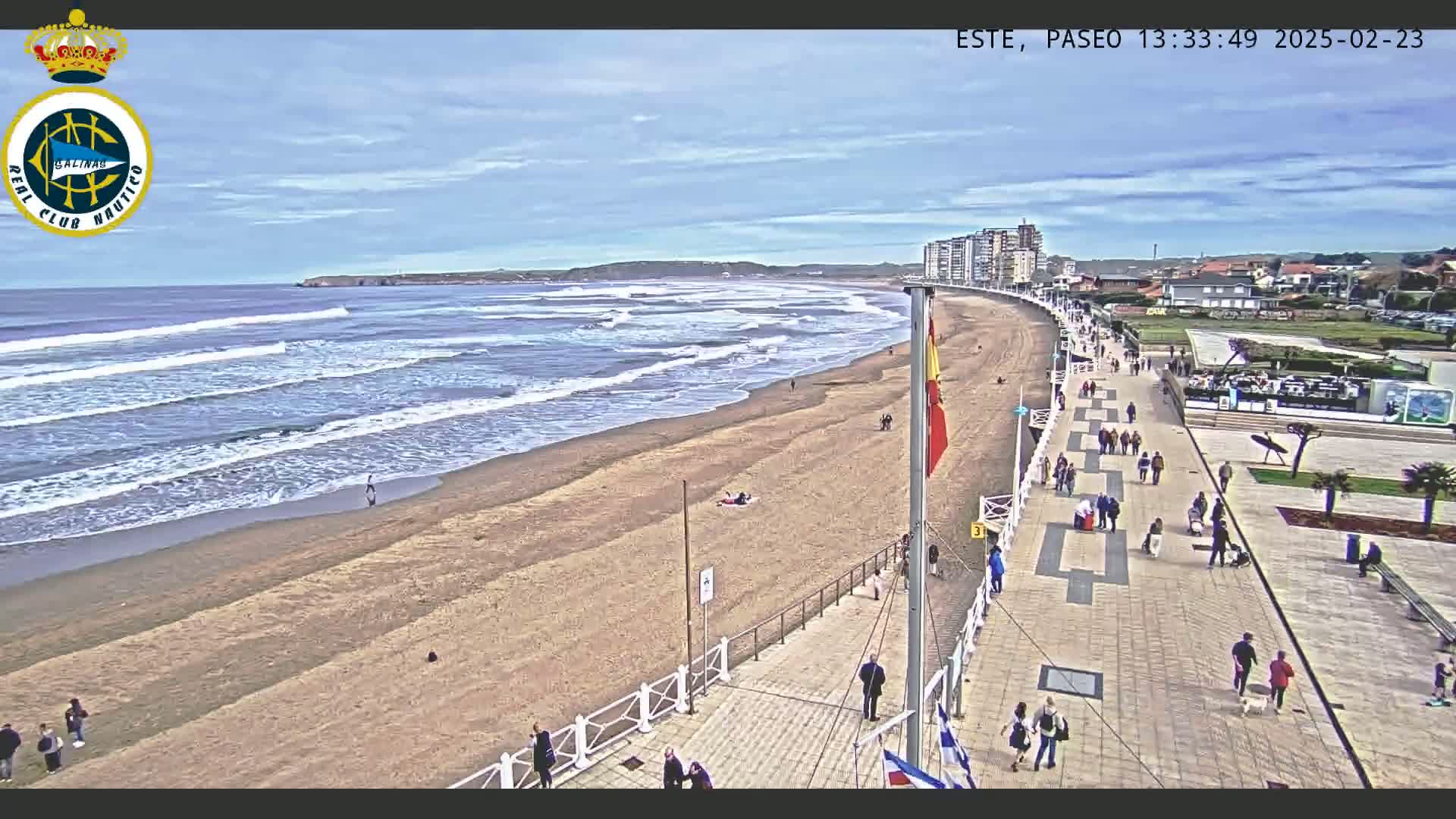 A sandy beach with moderate waves under a partly cloudy sky is bordered by a paved boardwalk with many people walking along it, and buildings are visible in the distance.