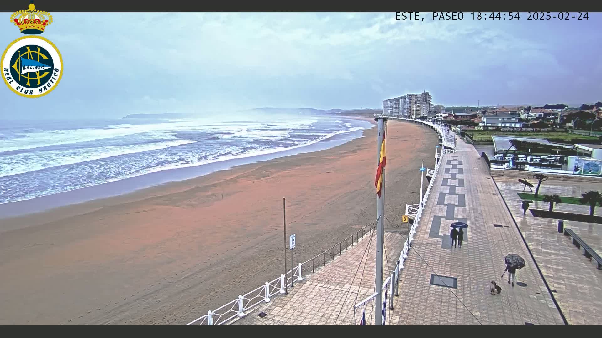 A sandy beach with waves rolling in under an overcast sky is bordered by a paved walkway with people walking under umbrellas, and buildings in the distance.