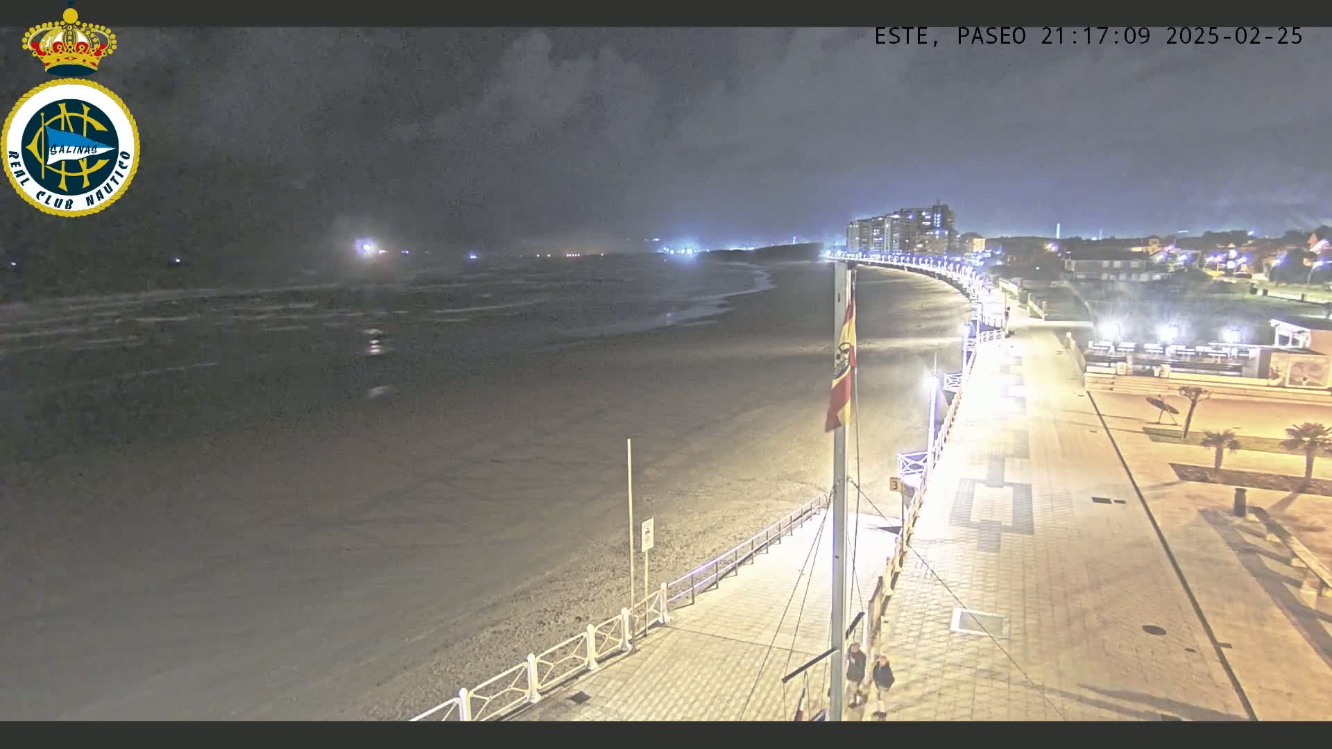 A nighttime view of a mostly empty beach with a paved walkway leading to buildings in the distance under a cloudy sky.