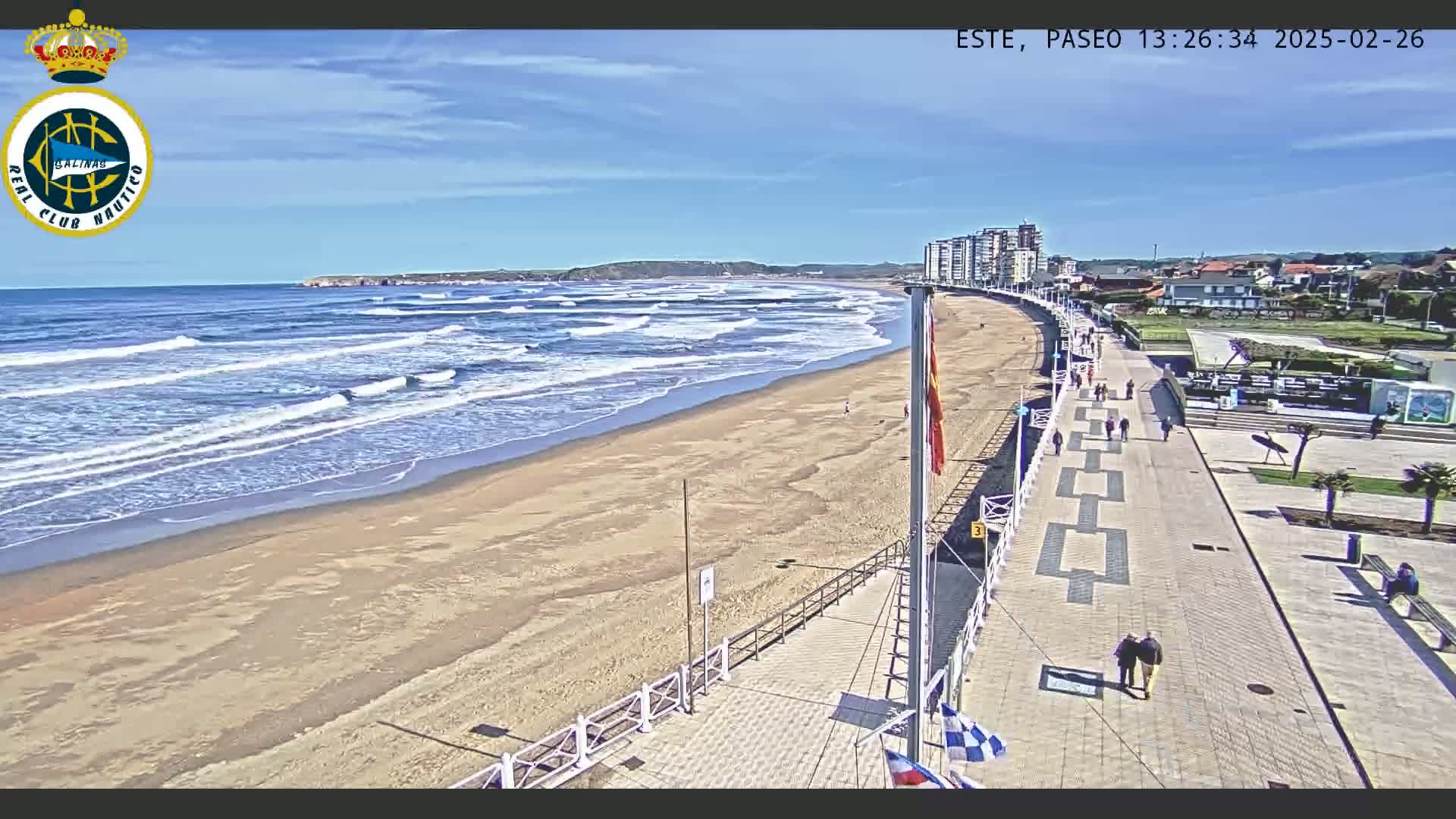 A sandy beach with gentle waves under a clear blue sky is bordered by a paved boardwalk with people walking, and buildings are visible in the distance.