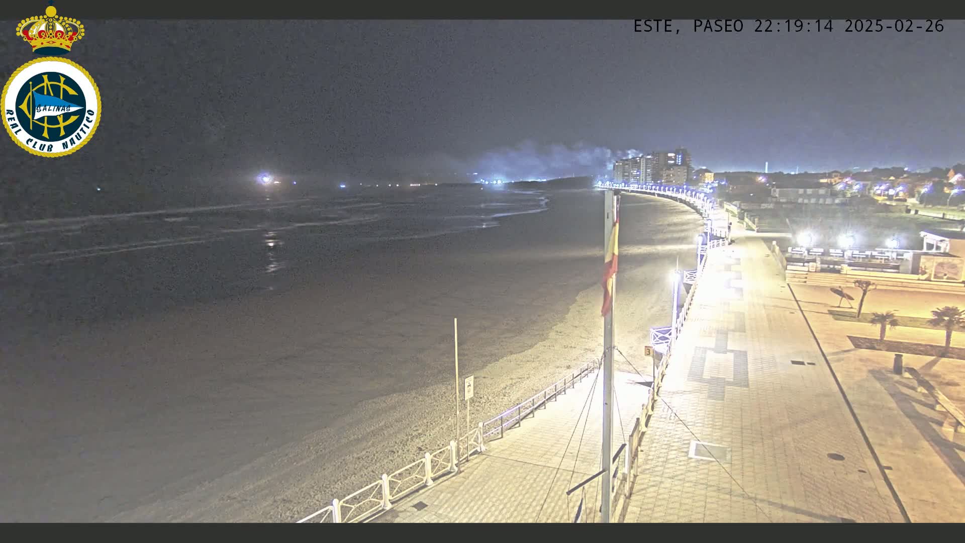 A nighttime beach scene shows a mostly empty sandy beach with gentle waves under a dark sky, next to a well-lit boardwalk with buildings in the distance.