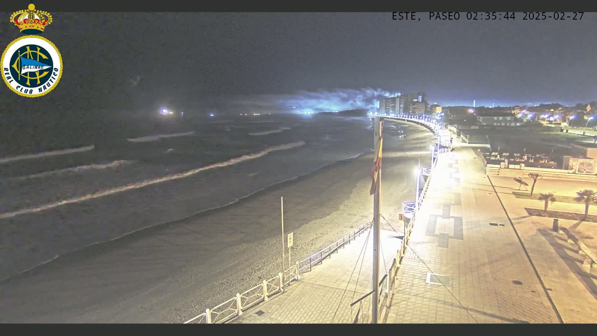 A nighttime view of a beach with gently rolling waves, a paved walkway, and buildings in the distance under a hazy sky.