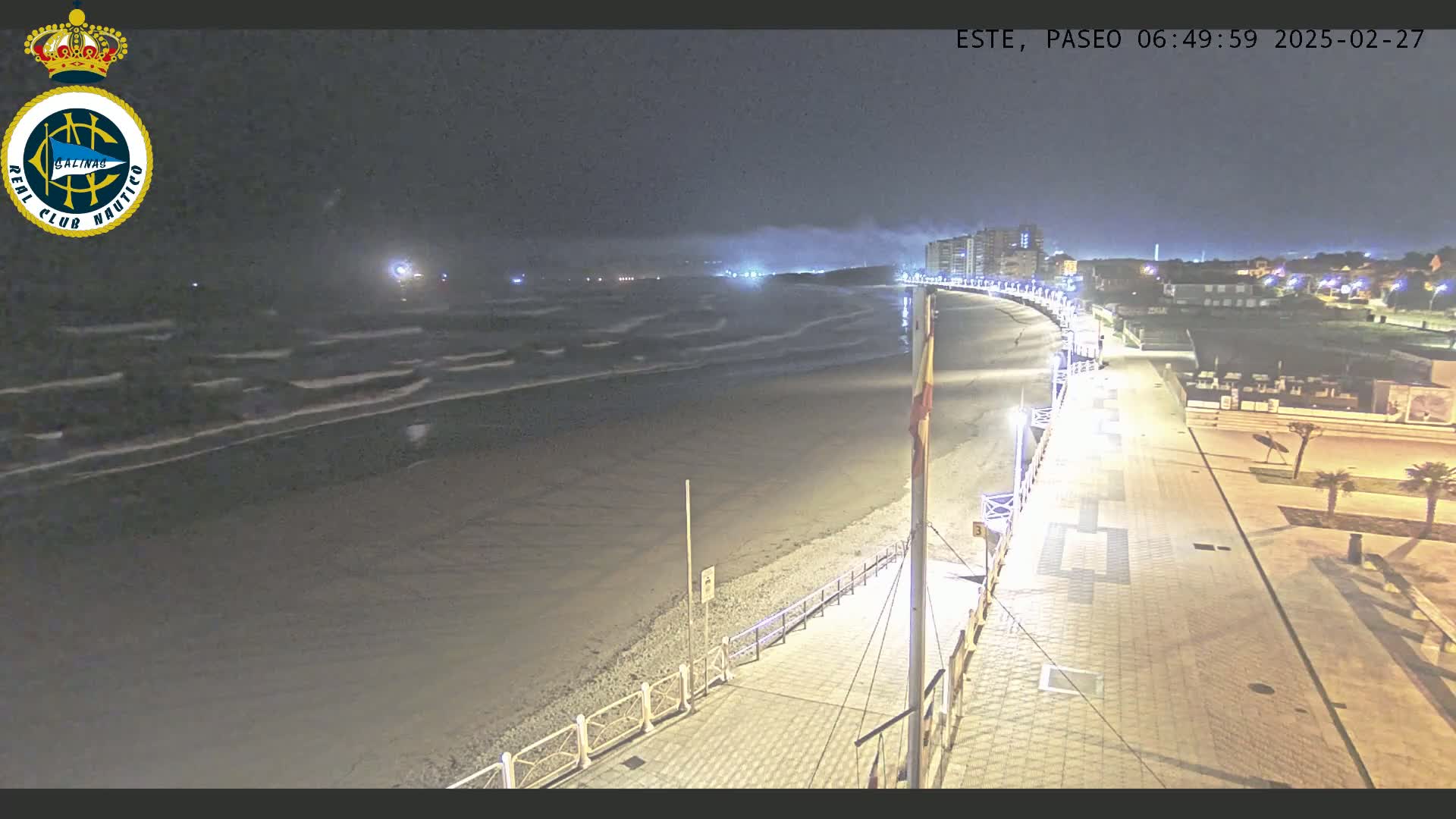 A nighttime view of a beach at low tide with moderate waves, showing a paved walkway lit by streetlights, leading to buildings in the distance under a cloudy sky.