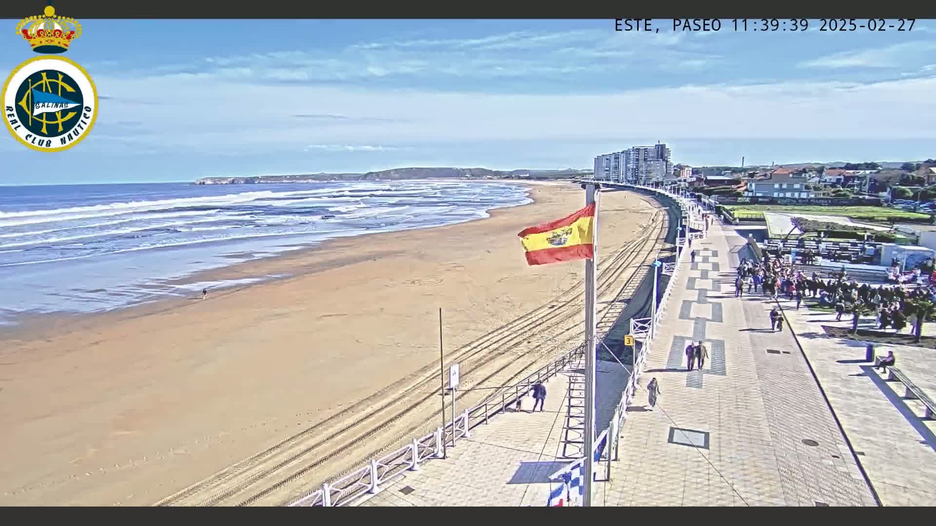 A sandy beach with gentle waves under a mostly sunny sky is bordered by a paved boardwalk with people walking, and buildings are visible in the background.