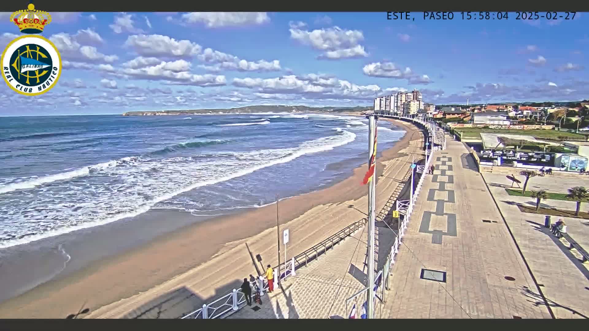 A sandy beach with gentle waves under a partly cloudy sky is bordered by a paved walkway leading to buildings and a small town.