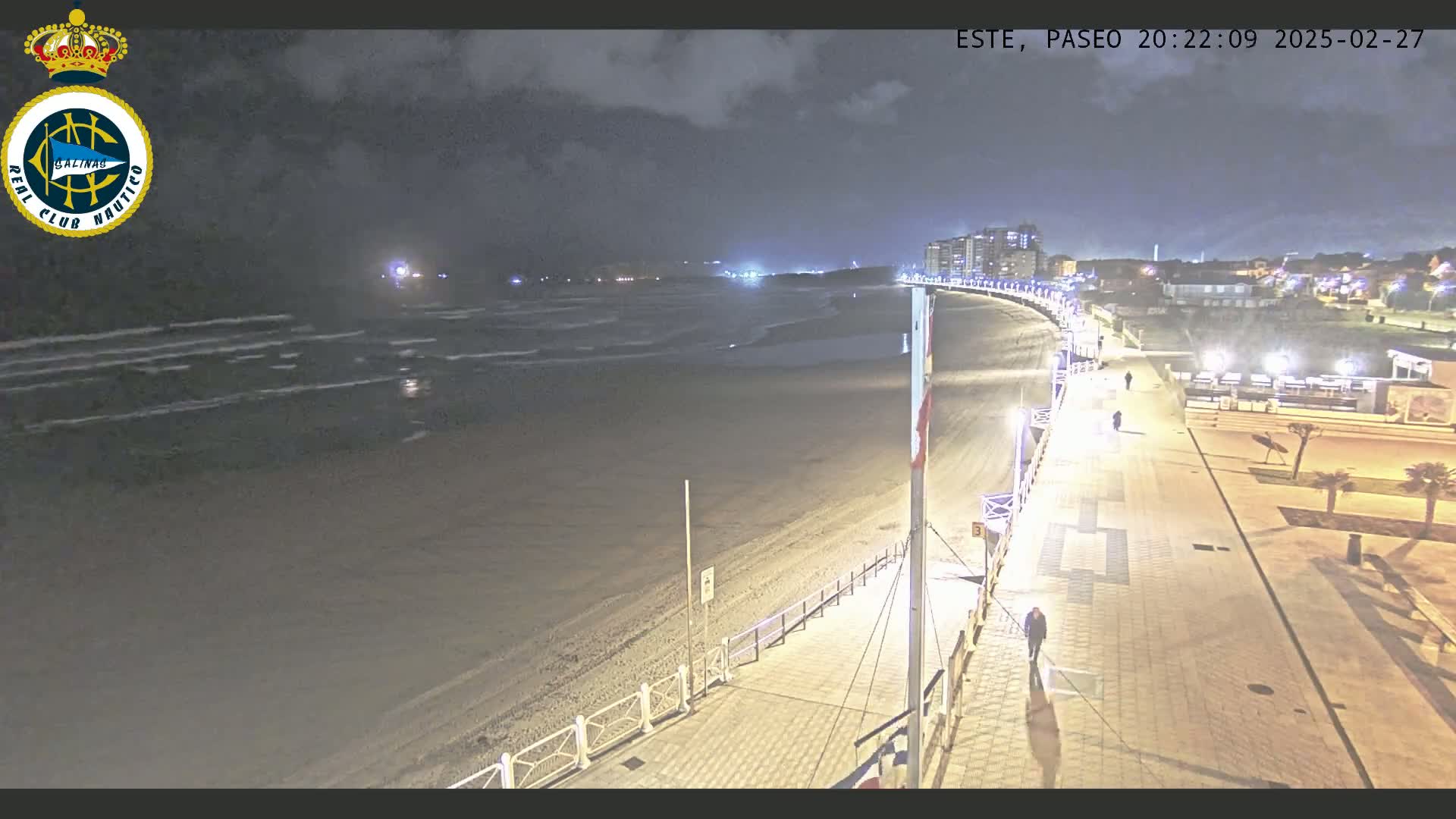 A nighttime view of a mostly empty beach with gentle waves under a cloudy sky, next to a brightly lit boardwalk with a few pedestrians.