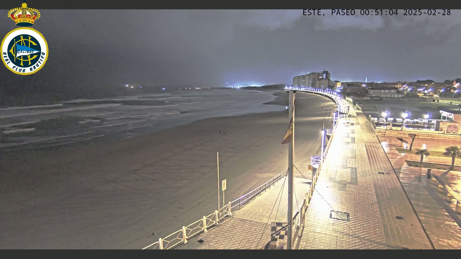 A nighttime view of a mostly empty beach with gentle waves, a paved boardwalk with lights, and buildings in the distance under a dark, cloudy sky.