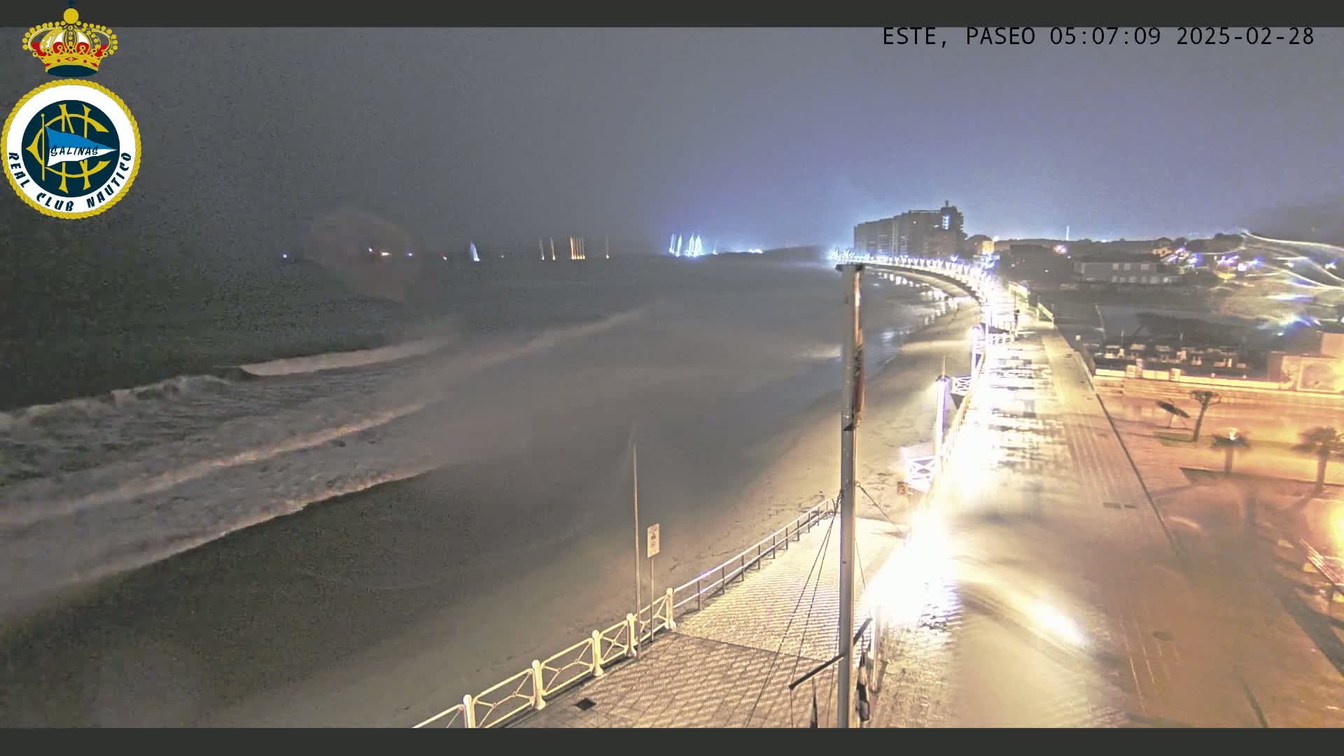 A nighttime coastal scene shows a beach with waves crashing ashore, illuminated by streetlights on a boardwalk next to buildings under a dark, stormy sky.