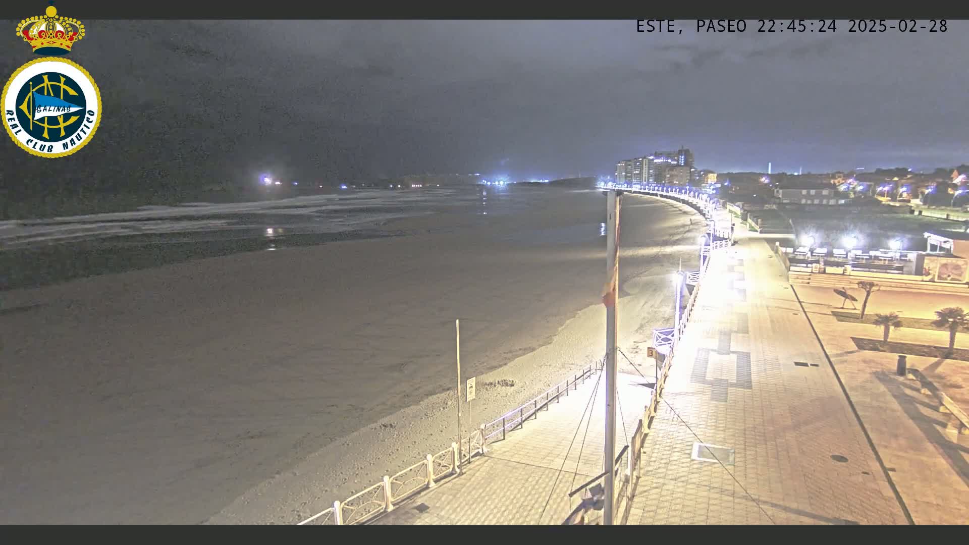 A nighttime view of a mostly empty beach with a paved walkway and buildings in the distance under a dark, cloudy sky.